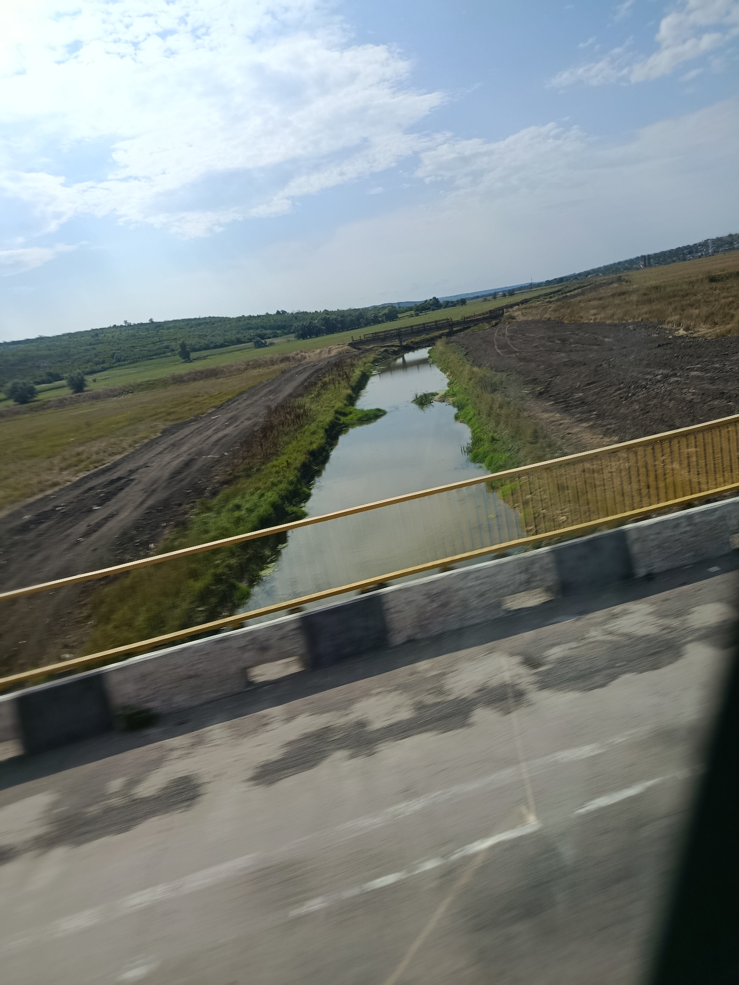 This image captures a landscape scene in Țipala, Moldova, viewed from a bridge. In the foreground, the surface of a road, likely asphalt, is visible with indistinct markings and shadows, suggesting motion blur from a vehicle. A concrete barrier with a yellow metal railing runs along the edge of the bridge. Beyond the bridge, a narrow waterway, possibly a canal or a small river, flows through the terrain. Its banks are lined with green vegetation and bare earth or disturbed soil on one side, indicating some form of construction or land management. The waterway is crossed by a simple wooden bridge in the mid-ground. The background consists of gently rolling hills covered with green fields and scattered trees. The sky is partly cloudy with a bright, clear blue expanse.