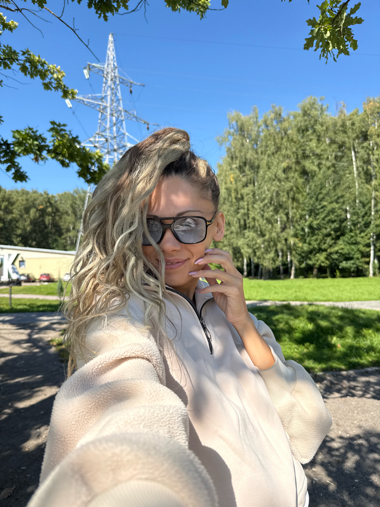 A mid-shot, outdoor selfie shows a woman in the city of Rusanovo, Russia, standing in front of a bright blue sky, trees, and a power transmission tower. The woman is wearing sunglasses and a light-colored, fuzzy jacket with a zipper. Her blonde hair is pulled up in a bun with some strands falling loose around her face. She is looking at the camera and touching her face with her hand. The background shows a green grassy area, trees, and a tall metal tower with power lines. In the distance, there is a yellow building and cars. The scene is brightly lit by sunlight, casting shadows on the ground.