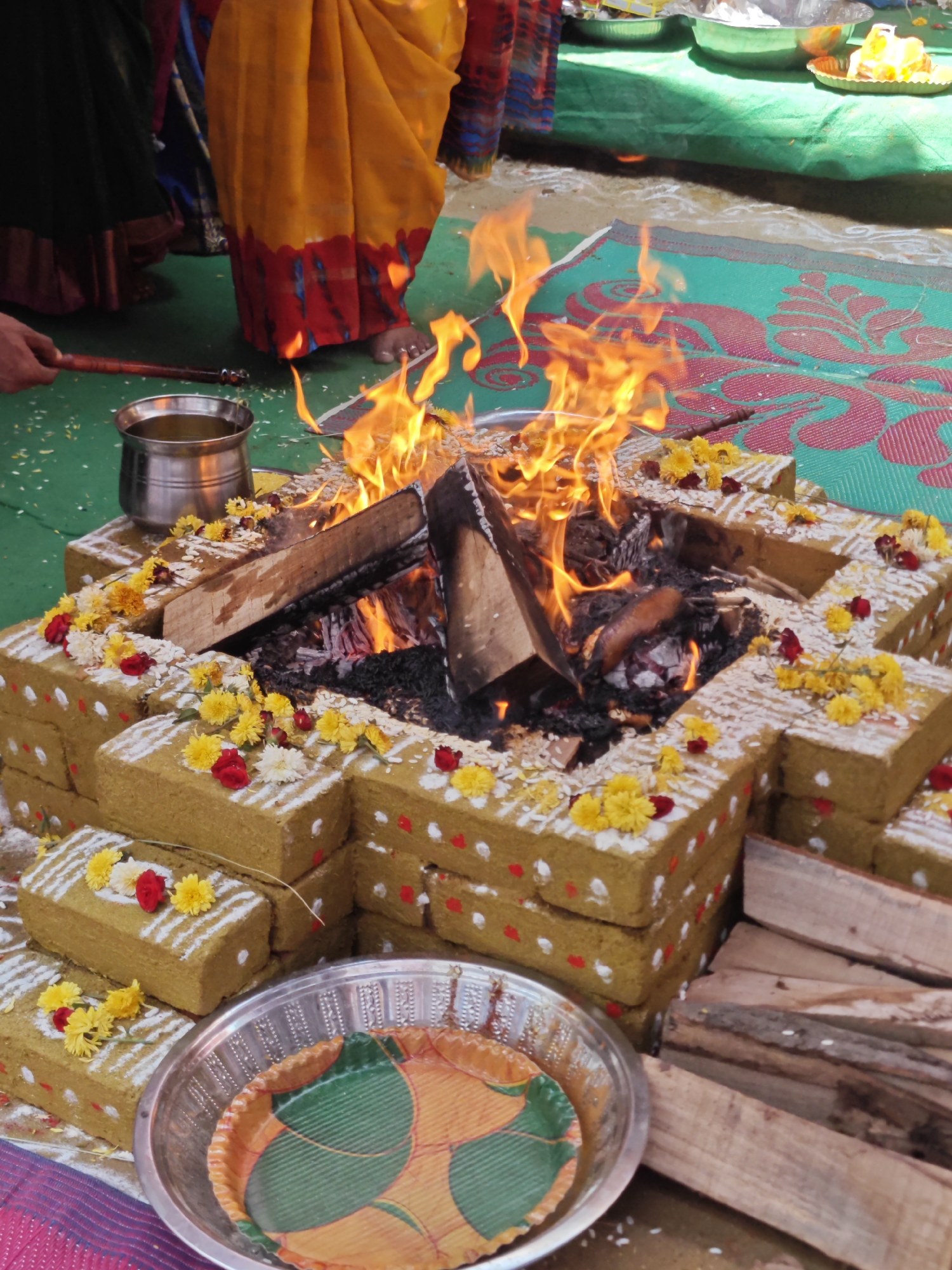 In Puttaparthi, India, a fire ritual is taking place, likely at a religious or cultural gathering. A brick structure, adorned with white powder markings, red and yellow flowers, and rice grains, forms the centerpiece. Logs are stacked to the right, ready for the fire. A silver pot containing liquid sits to the left, with a person's hand holding a wooden stick near it, possibly for tending the fire. Flames erupt from the center of the structure, illuminating the scene. In the background, people are partially visible, dressed in traditional Indian attire, including a vibrant yellow and orange saree. The ground is covered by a green mat with a pink patterned design, and there are scattered rice grains.