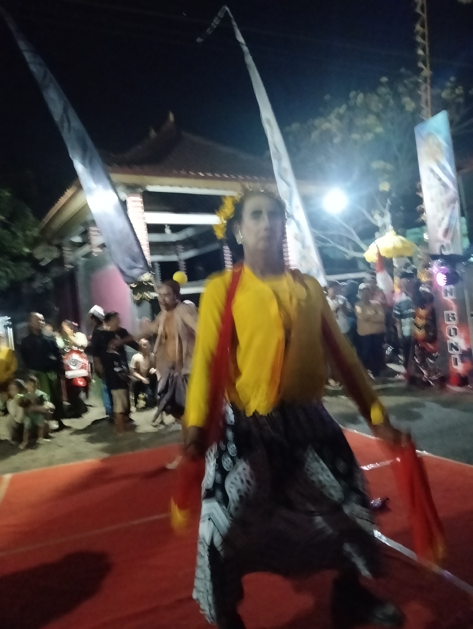 A blurry, night-time photograph shows a crowd of people gathered on a street in Sumbersewu, Indonesia. The main subject, a performer wearing a yellow top and a patterned skirt, is in the foreground, with their body angled away from the camera. They appear to be dancing or performing a ritual, holding a red cloth. Behind the performer, a red carpet is laid out on the ground. The background features a traditional building with an ornate roof, along with several tall flags and banners. Several individuals are visible in the crowd, some standing, others seated, and a red scooter is parked to the left. The overall atmosphere suggests a local cultural event or celebration.
