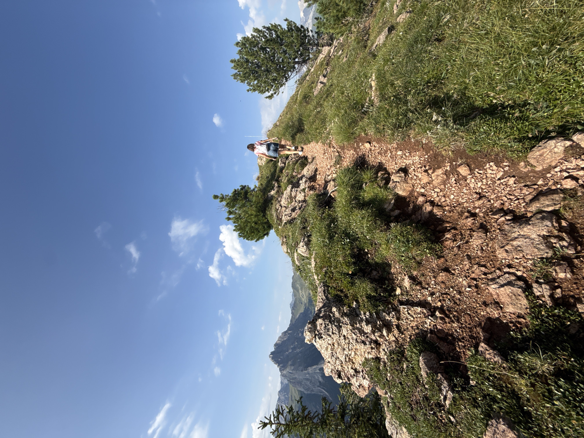 A woman with dark hair, wearing a pink shirt and denim shorts, walks up a rocky, winding mountain trail. She carries a white backpack. The trail is lined with green grass and scattered rocks. In the background, large, rugged mountains are visible under a bright blue sky with scattered white clouds. A few evergreen trees are also present on the mountainside. The image depicts a hiking scene in a mountainous, natural environment. The location is a mountain landscape near Canazei, Italy.