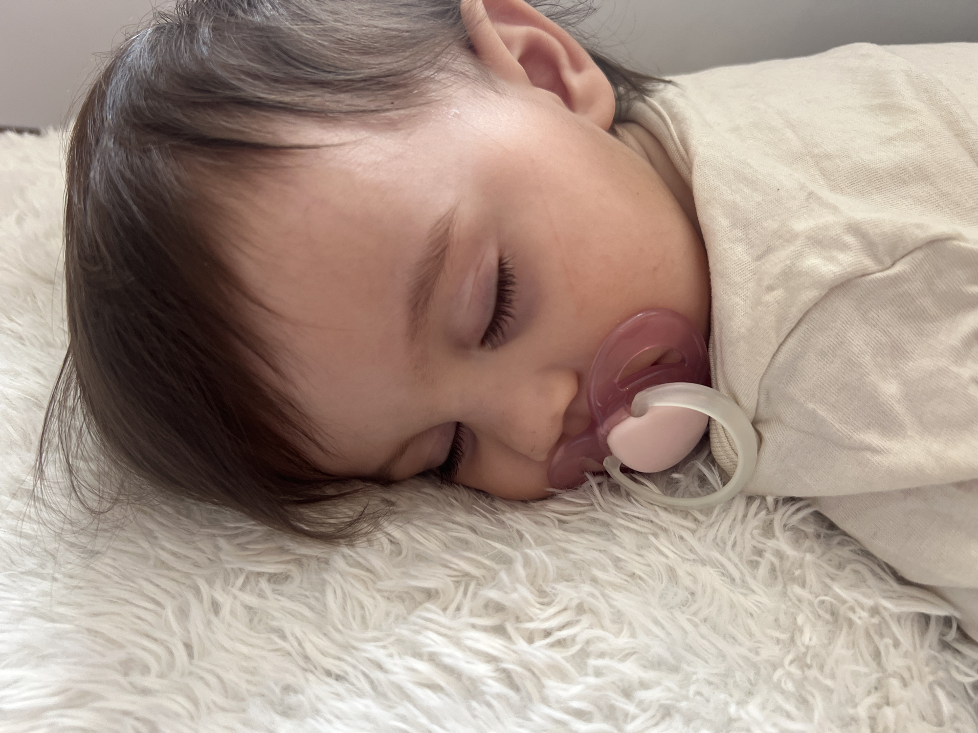A baby with dark hair and closed eyes is lying on a fluffy white surface. The baby has a pink and white pacifier in its mouth. The baby is wearing a light-colored shirt. The setting is indoors. The location is unspecified in the city of Elena, Bulgaria.