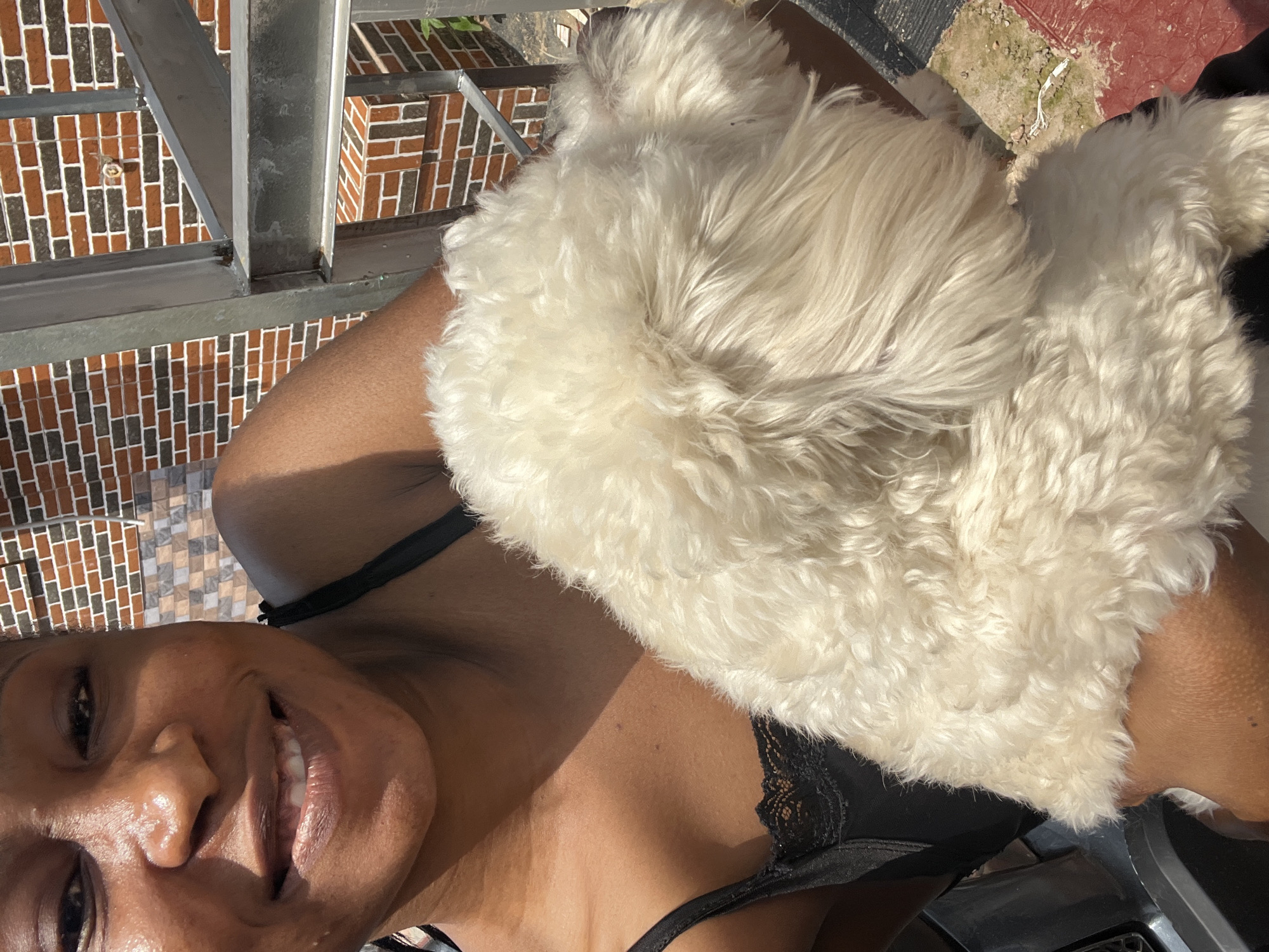 A woman with dark skin and a bright smile is captured in a close-up, horizontal selfie, her head tilted to the side. She's wearing a black lace-trimmed bra, and a fluffy white dog is nestled against her neck and shoulder, obscuring part of her face. Sunlight highlights her skin, creating a warm glow. In the background, a metal railing and a brick wall with a patterned tile facade suggest an outdoor setting, possibly a balcony or patio. The scene is bathed in bright sunlight, indicating it's daytime. The woman's joyful expression conveys a sense of happiness and affection, likely towards the dog. The overall mood is relaxed and personal.
