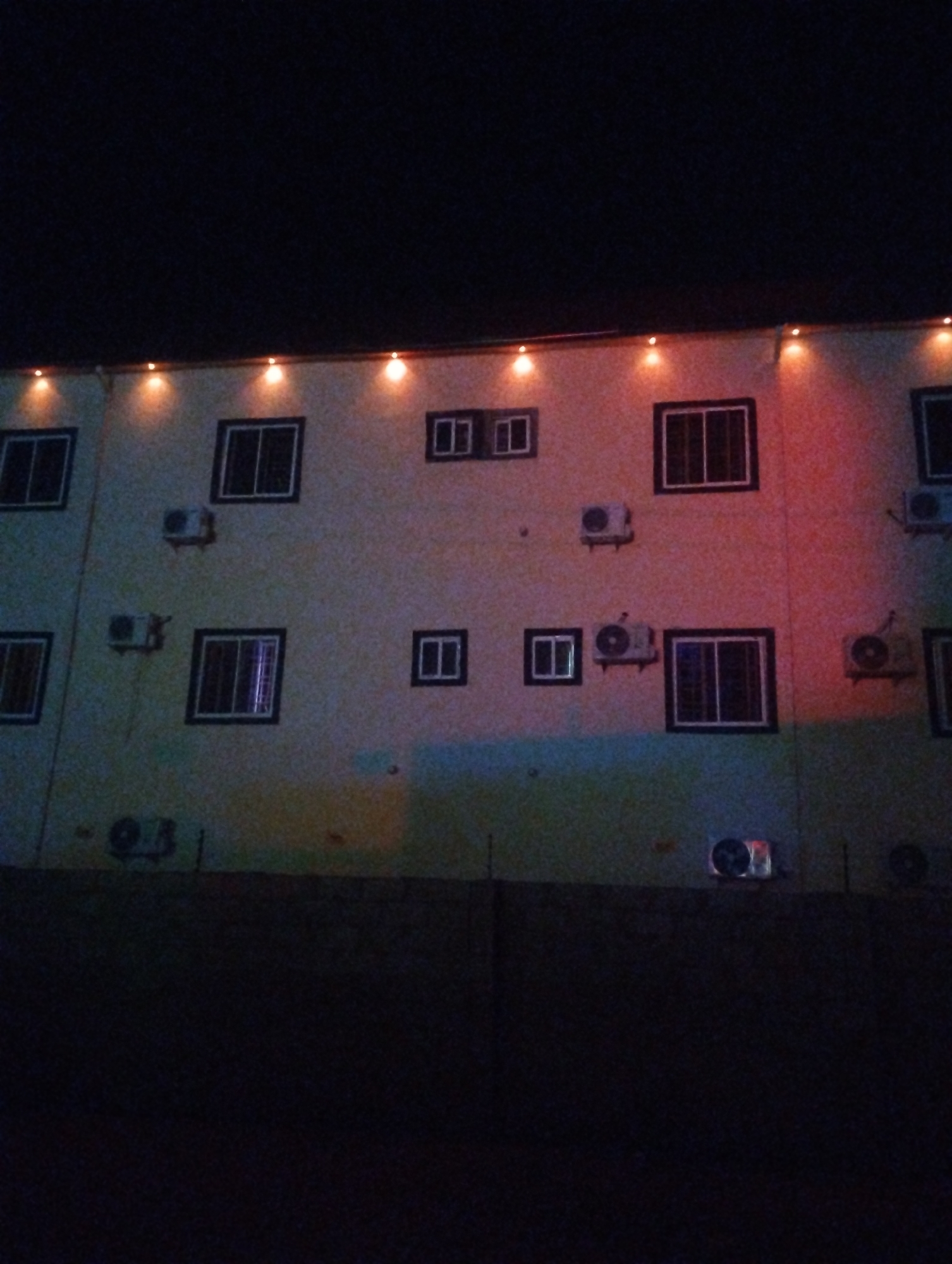 The image depicts the exterior of a building in Tumfure, Nigeria at night. The building is multi-story with a beige facade, dark-framed windows, and numerous air conditioning units mounted on the walls. A string of lights adorns the top edge of the building. The ground in front of the building is dark and obscuring. There is no evidence of human presence or activity.