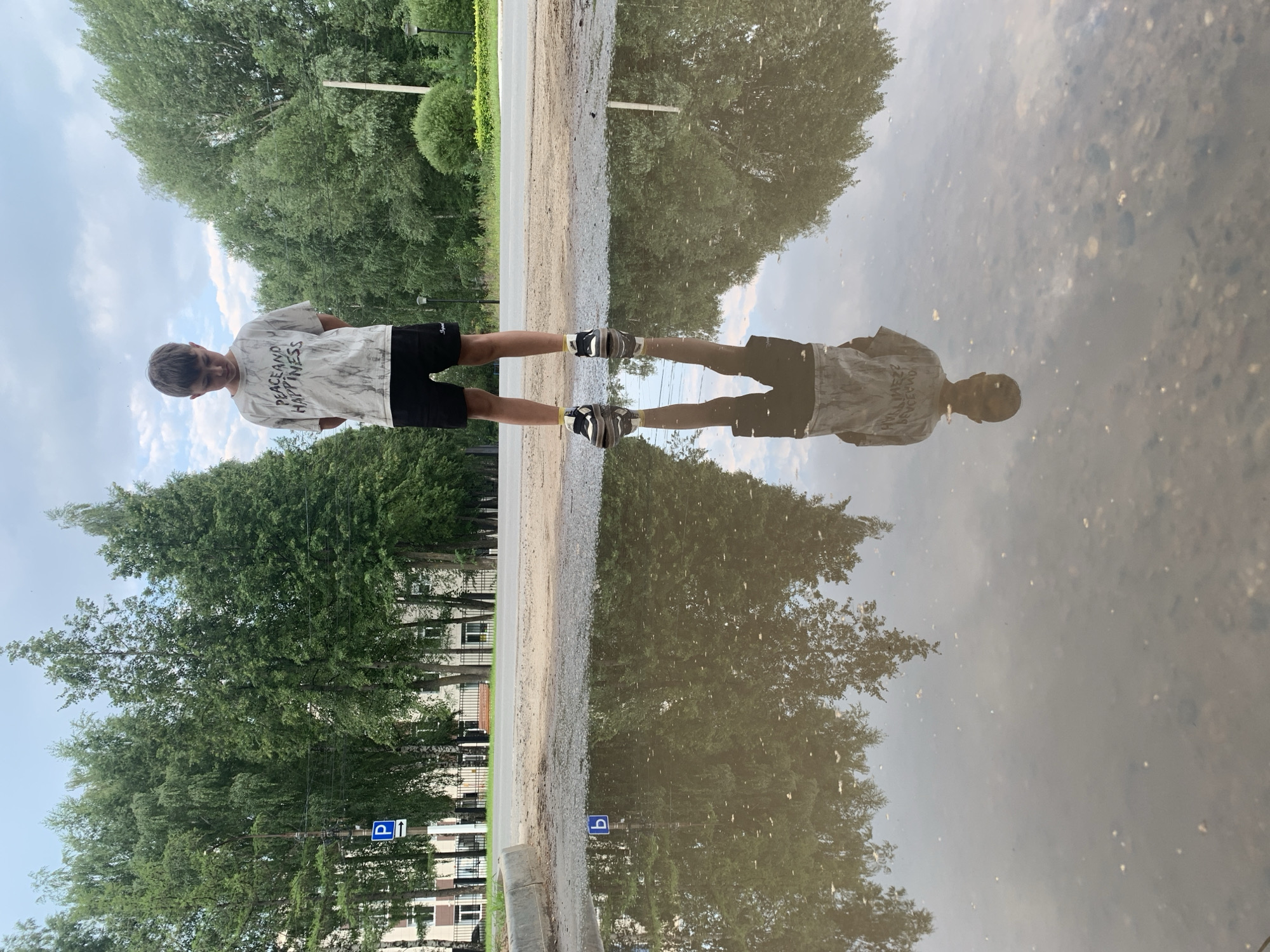 A young boy stands in the middle of a large puddle on a paved surface, with trees and buildings in the background. The boy is wearing a grey t-shirt with the words "PEACE AND HAPPINESS" printed on it in black, and black shorts. He is looking down at his reflection in the puddle. The puddle is reflecting the sky, trees, and buildings, as well as the boy's reflection. The sky is cloudy. There is a blue parking sign with a white "P" and an arrow pointing to the left, visible both directly and reflected in the puddle. The scene appears to be in the city of Chaikovsky, Russia, given the context.