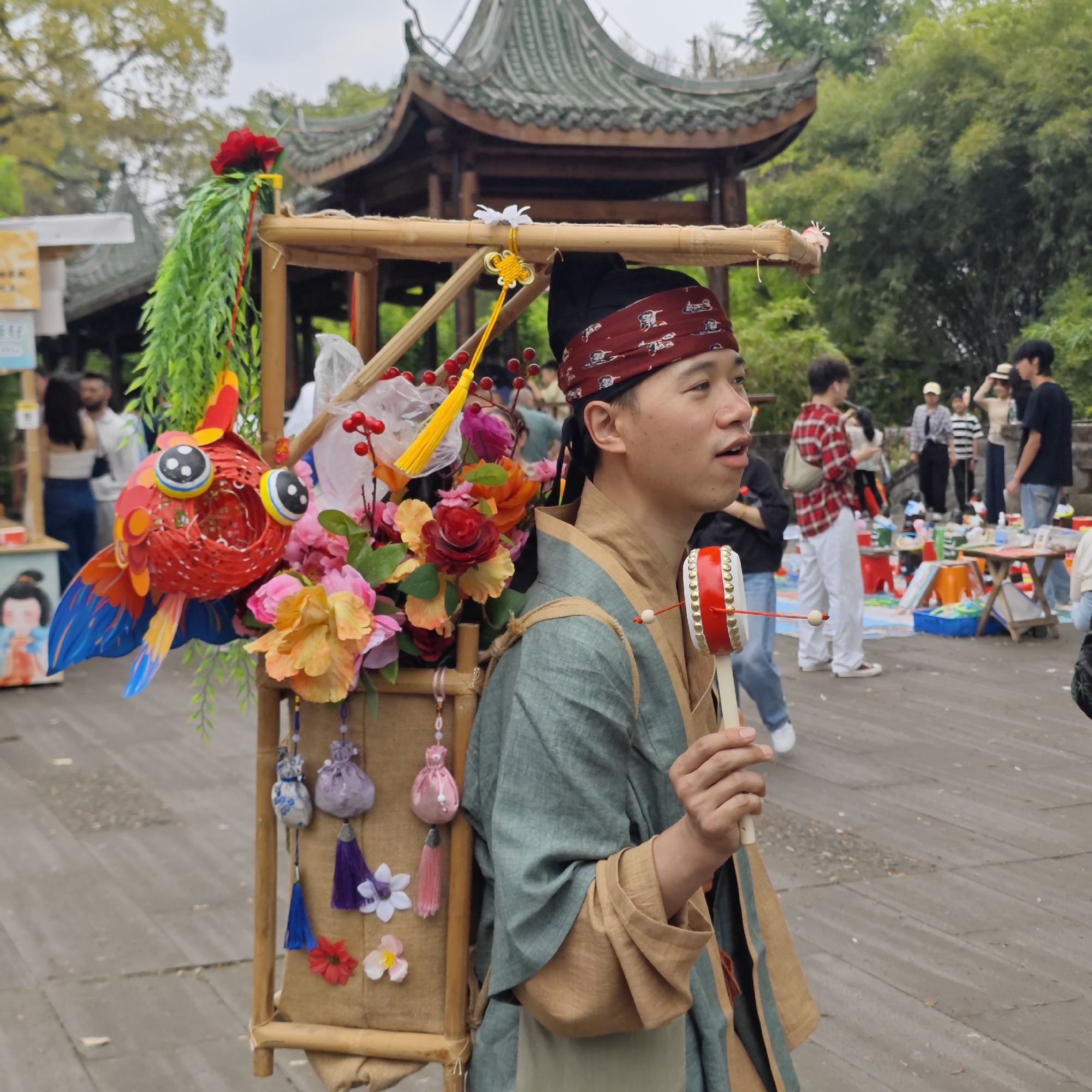The image captures a man dressed in traditional Chinese attire, standing in what appears to be a marketplace within a park or garden setting. He wears a green robe with tan accents and a red headband. Strapped to his back is a bamboo frame adorned with colorful decorations including a large paper fish, artificial flowers, and small fabric pouches with tassels. In his hand, he holds a small, double-sided drum.
The backdrop includes a traditional Chinese gazebo with a multi-tiered roof, suggesting a historic or cultural park. Several people are in the background, some gathered around what looks like vendor stalls, implying a market or fair is taking place. The setting is likely Yuzhong District, China, given the traditional architecture and cultural elements evident in the scene. The weather appears overcast.
