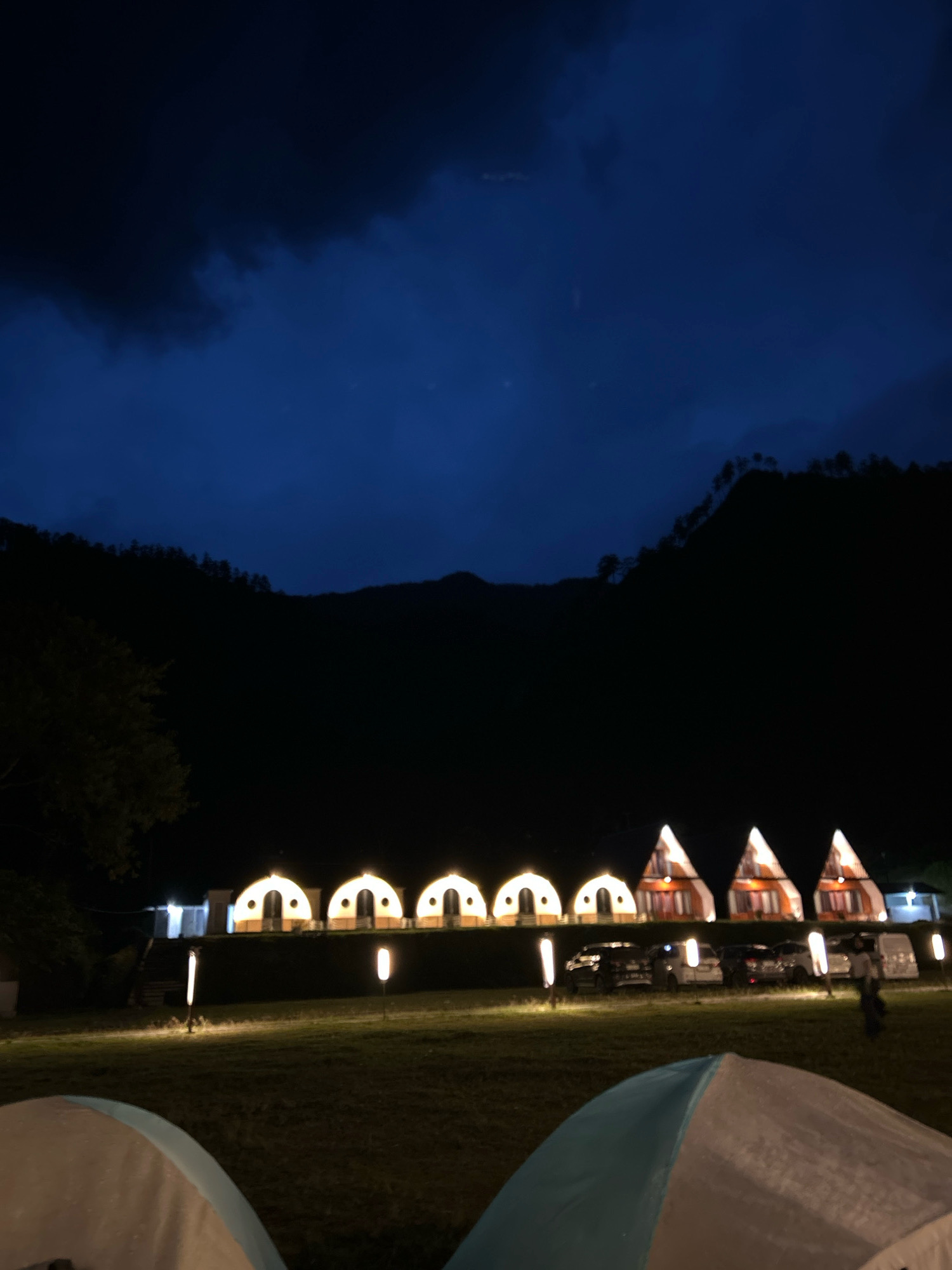 At night in Merodot, Indonesia, a campsite is illuminated by artificial lights. In the foreground, two tents are partially visible, their fabric reflecting the ambient light. Behind them, a row of small, dome-shaped structures and triangular cabins are lit from within, creating a warm glow. Several vehicles are parked in front of the cabins, their headlights and taillights adding to the illumination. Tall, slender lampposts are placed strategically throughout the campsite, casting pools of light on the grassy ground. In the background, dark mountains rise against a deep blue, cloudy sky, their silhouettes adding a sense of depth to the scene. A single person can be seen walking near the vehicles on the right side of the image. The overall atmosphere is peaceful and inviting, suggesting a place for rest and relaxation.