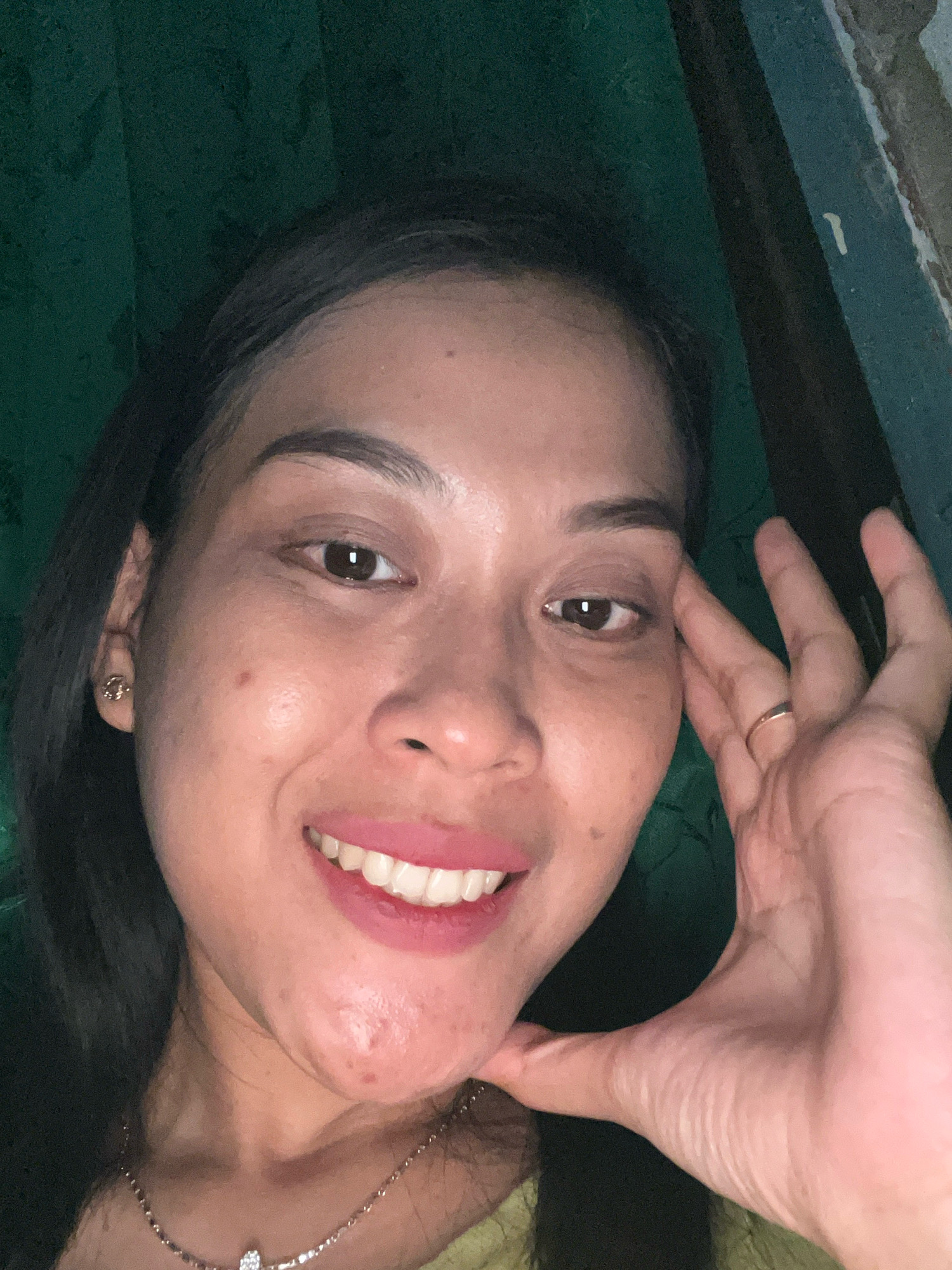 A close-up, eye-level, indoor, selfie-style portrait shows a young woman with dark hair and brown eyes. She is smiling, exposing her teeth, and her right hand is resting on her cheek. She is wearing a gold stud earring and a thin gold ring on her left hand. A necklace with a jeweled pendant is visible around her neck. The background is a dark green fabric or curtain, with a dark, possibly wooden, structure on the right side of the frame. The lighting is dim, creating a warm glow on the woman's face. The scene appears to be set in a residential environment within Kecamatan Pati, Indonesia.