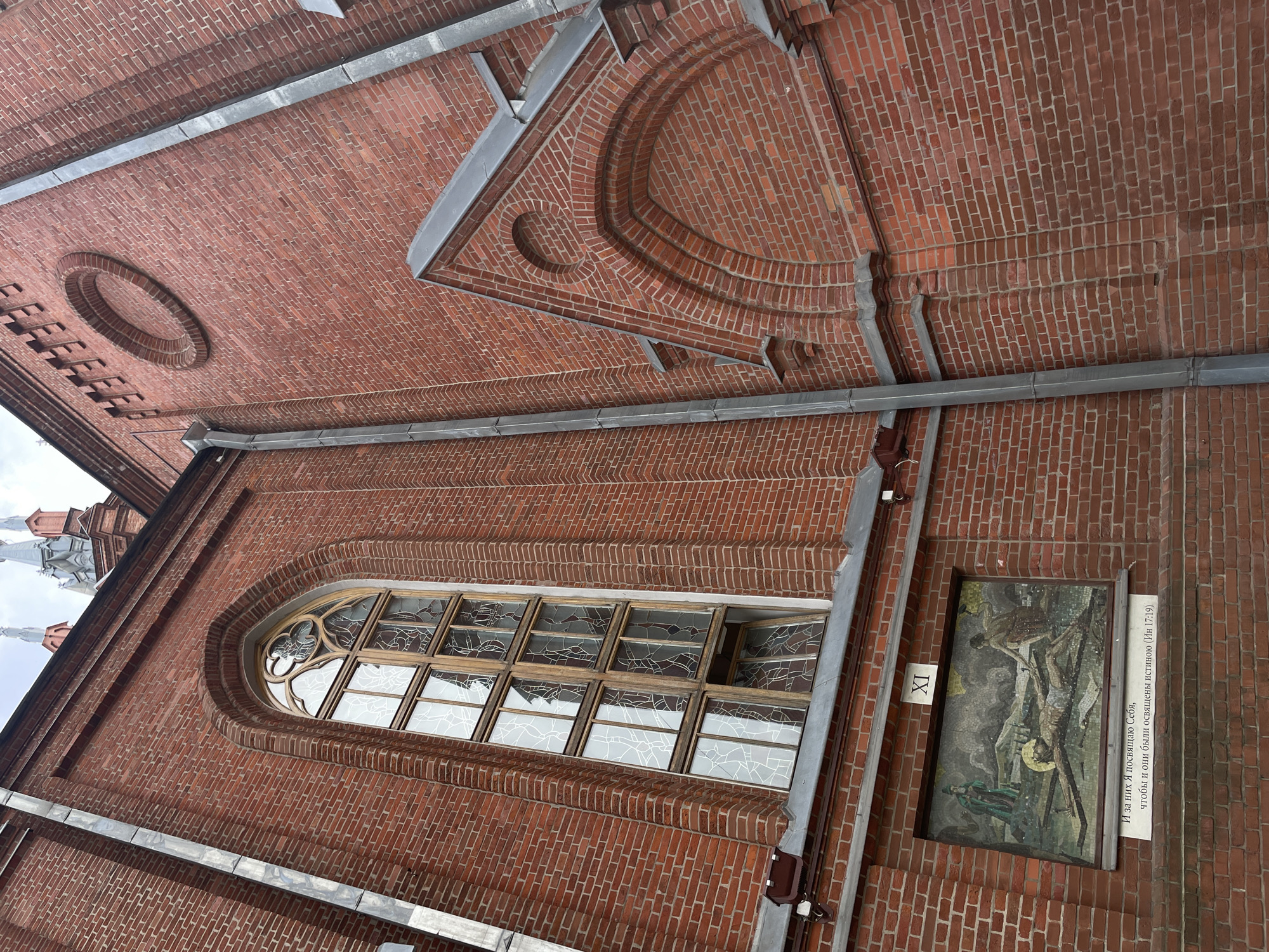 This image shows a close-up, worm's-eye view of the exterior of a red brick building, likely a church given the architectural details and the mosaic artwork. The building features a large, arched window with ornate stained glass panes. Above the window, decorative brickwork creates a circular indentation within a triangular recess. Along the brick wall, a metal gutter runs horizontally, and below the window, a mosaic artwork is set into the wall. To the right of the mosaic, a plaque contains text in Cyrillic. The mosaic depicts a scene with several figures, one of whom appears to be crucified. The text on the plaque, which is partially visible, mentions a religious verse. The sky is overcast, suggesting a cloudy day. The overall impression is one of historical and religious architecture, with a focus on the craftsmanship of the brickwork and the artwork. The scene is static, presenting a detail of the building's facade.