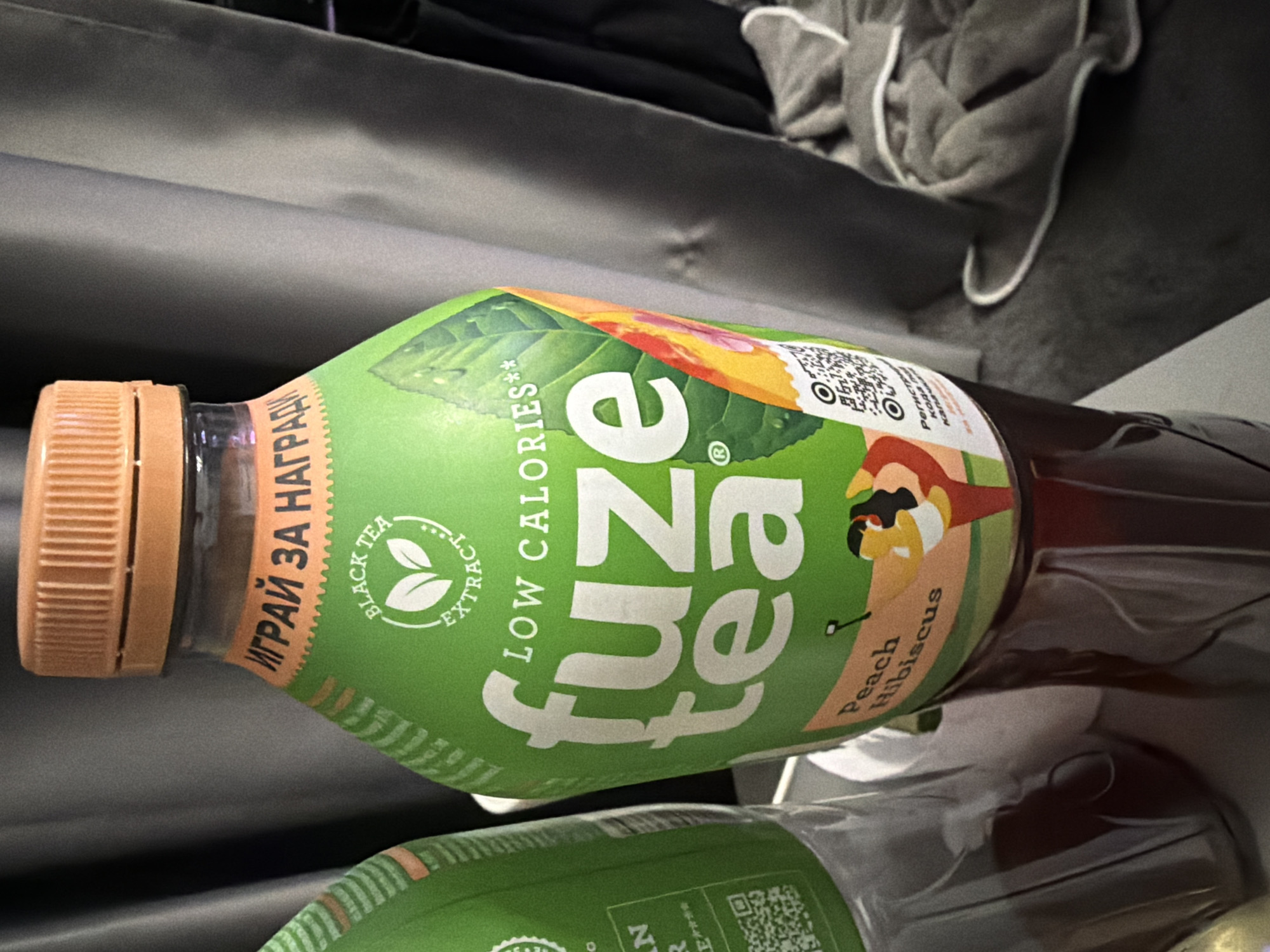 The image shows two bottles of Fuze Tea, one in focus and another slightly out of focus to the left. The bottle in focus is bright green with white text and graphics. It features the "Fuze Tea" logo prominently in large white letters. Above the logo, in orange text with a scalloped edge, is Russian text that translates to "PLAY FOR A PRIZE". Below the logo, there's text indicating "LOW CALORIES" and a graphic of leaves. The flavor is identified as "Peach Hibiscus" with a small illustration of a woman in athletic wear. A QR code is also visible on the bottle. The bottles are placed on a reflective surface, possibly a table or counter. In the background, dark grey fabric drapes, and to the right, a grey towel with white trim is draped over something unseen. The lighting is dim, creating shadows and highlights. The image appears to be taken indoors. There is no indication of the city of Ruse, Bulgaria.