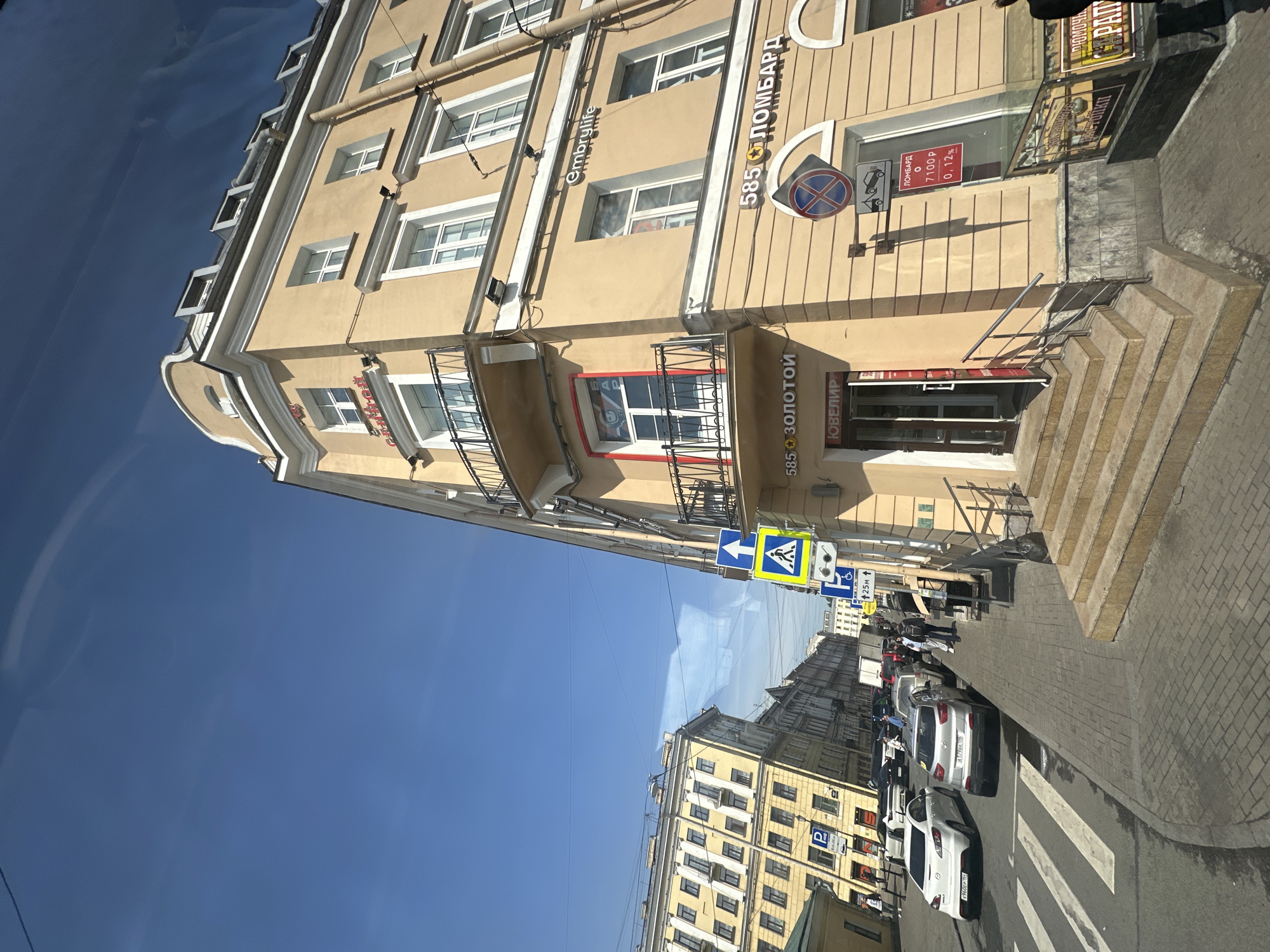 The image presents a street scene in Saint Petersburg, Russia. It captures a portion of a multi-story building with a beige facade, detailed window frames, and signage written in Cyrillic script. A sign for "585 Золотой" (585 Gold) is visible near the entrance. Additional signs indicate a pawn shop ("Ломбард") and other commercial activities.
On the street, there are several parked cars and a pedestrian crosswalk. Sidewalk infrastructure, including crosswalk signs, are evident. Other buildings are seen down the street. The sky is blue and clear. The scene is viewed from a slightly oblique angle.