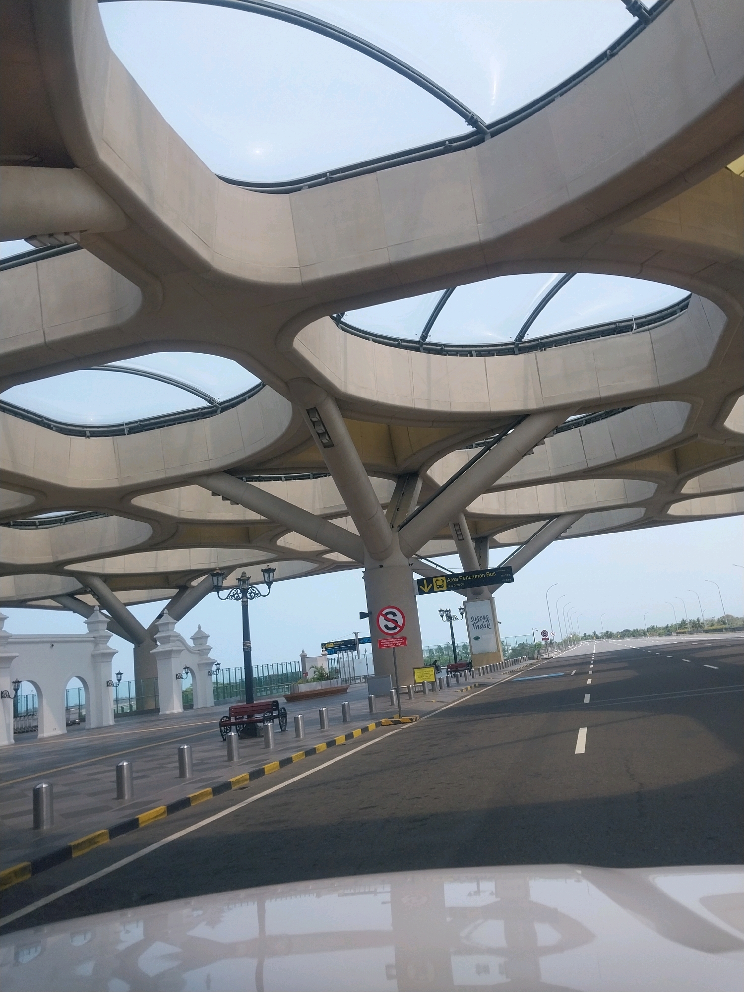 The image depicts the exterior of an airport terminal in Palihan, Indonesia, likely the Yogyakarta International Airport. The focal point is the building's distinctive canopy, a modern architectural structure characterized by a series of interconnected, curvilinear concrete beams forming an organic, honeycomb-like pattern. Large, oval-shaped openings within the canopy reveal a clear sky, suggesting a daytime scene.
The area directly beneath the canopy appears to be an open-air walkway or drop-off zone. Several bollards are arranged along the edge of the road, and a red bench with decorative metalwork is visible to the left. White arched structures, possibly entrances or decorative elements, line the building's facade. A lamppost with multiple ornate lights stands near the bench.
To the right, a wide asphalt road stretches into the distance, marked with white dashed lines indicating lanes. The road appears to be part of the airport's access or departure routes. Various road signs are present; one prominently displays a "No Entry" symbol, and another, in Indonesian, directs to "Area Penurunan Bus" (Bus Drop-off Area) and "Bus -- -- -- -- --" likely indicating bus routes or bay numbers. A smaller sign on a pole reads "Sugeng Tindak," a Javanese phrase for "Farewell" or "Safe Journey."
The overall environment suggests a clean, well-maintained public space, typical of a modern transportation hub. The lighting indicates it is likely morning or late afternoon, given the angle of the shadows and the brightness of the sky. No people are clearly visible in the image, contributing to a sense of quietude. The reflection of the sky and canopy on the white surface in the foreground suggests the viewer is inside a vehicle.