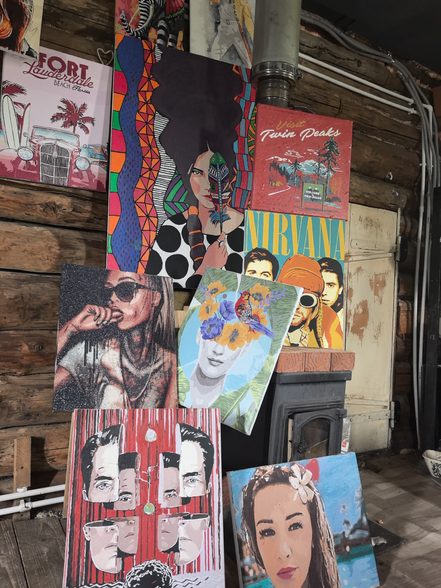The image shows a collection of paintings stacked and leaning against a log wall in what appears to be an interior space in Noviy, Russia. The walls are constructed of wooden logs. The collection includes paintings that are of different styles: pop art, portraiture, and vintage travel advertisements. "Fort Lauderdale" and "Twin Peaks" can be read in the paintings. One painting features a collage of human faces. In the background, there is a metal pipe and a closed door. The space feels cluttered and artistically oriented. A small stove appears behind several of the paintings.