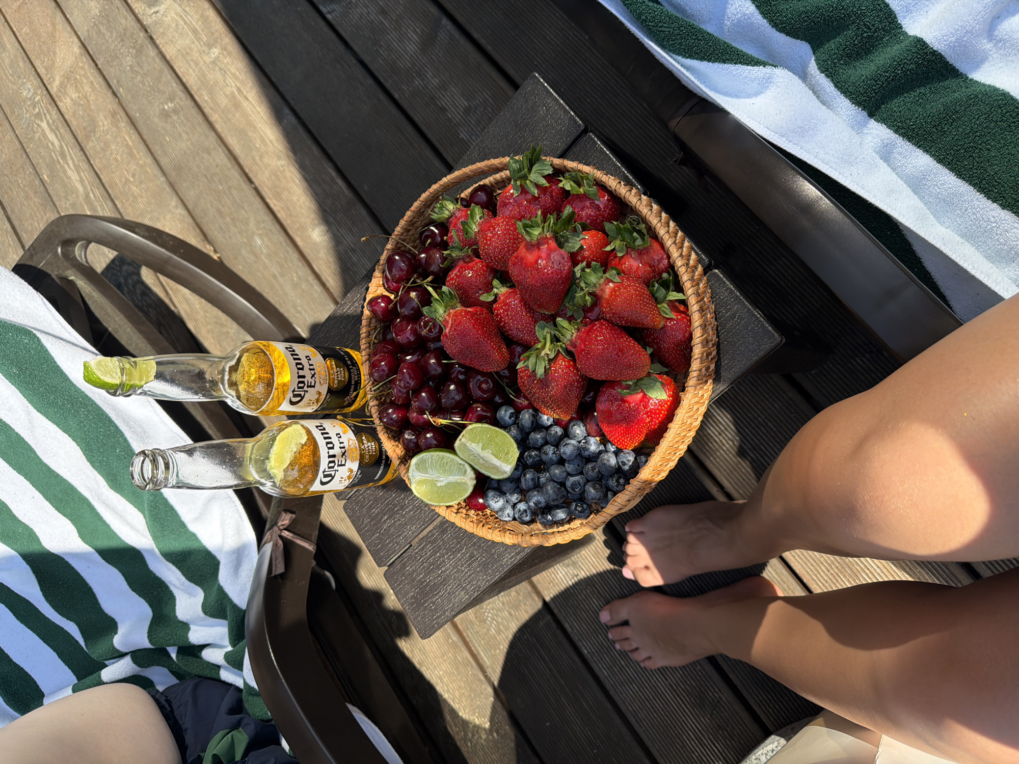 The scene depicts an outdoor leisure setting in Mansky, Russia, under bright daylight. A person's tanned bare legs and feet are visible in the foreground, resting on dark wooden decking, with distinct shadows indicating direct sunlight. On the left side, the upper thigh and hip area of a person, wearing dark shorts, are partially visible, seated on a dark brown sun lounger draped with a white towel featuring broad green stripes.
A small, dark brown rectangular side table is positioned on the decking. On this table rests a round, woven wicker basket brimming with fresh produce: numerous vibrant red strawberries with green caps, a substantial quantity of dark blue blueberries, and a pile of dark red cherries with stems. Two halves of a fresh green lime are placed atop the fruit in the basket. Adjacent to the basket, two clear glass bottles of "Corona Extra" beer stand upright, each with a fresh lime wedge inserted into its neck. A second sun lounger, also covered by a green and white striped towel, is partially visible on the lower right side of the frame. The wooden decking surface displays a consistent dark brown hue with visible grain.