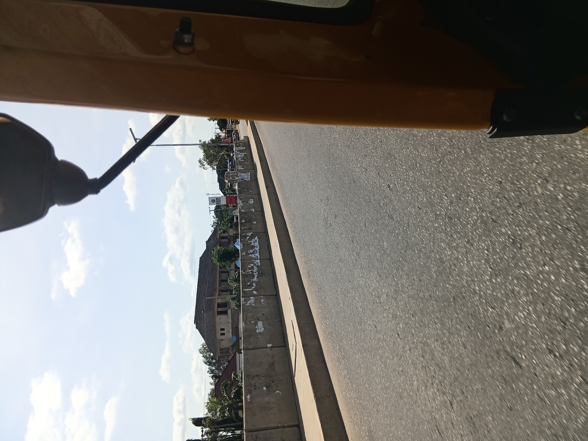 This image captures a view from the perspective of someone traveling in a yellow auto-rickshaw in Jalingo, Nigeria. The scene is dominated by a road, with a concrete barrier along its side. Beyond the barrier, there is a building with a dark, tiled roof and several windows. Trees and other smaller buildings are visible in the background. The sky is light blue with scattered white clouds. The right side of the image shows part of the auto-rickshaw, including its yellow bodywork and a black component attached to it. The road surface is asphalt, and appears to be in moderate condition with some white speckling.