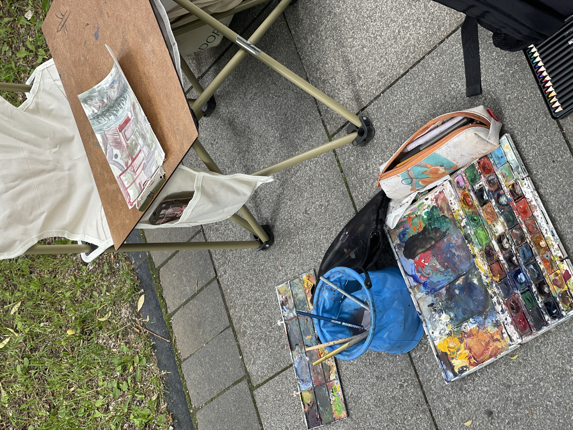 An outdoor scene shows an artist's setup for watercolor painting on a paved area next to a grassy patch. A portable folding table with a wooden surface holds a watercolor painting of a red building and trees. To the left of the table, on the ground, are two paint palettes overflowing with various colors, a blue bucket containing brushes and water, and a pencil case adorned with a fox. A black backpack is visible to the right. The overall impression is of an artist engaged in their craft in a public space.