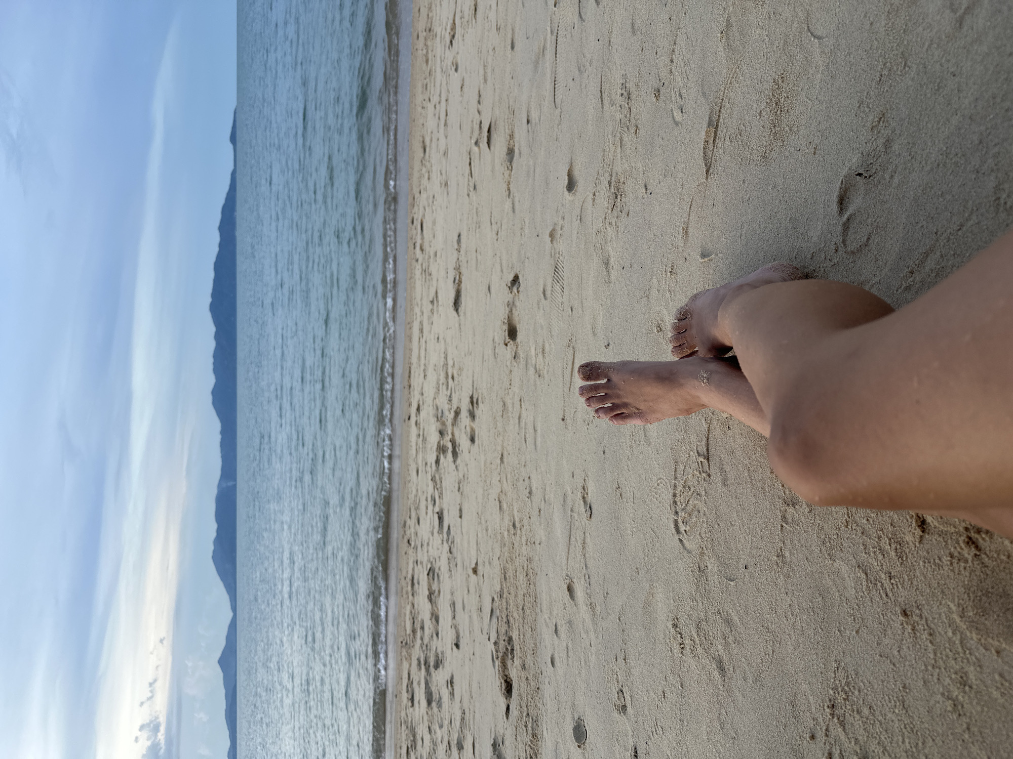 A first-person perspective shot of a person's legs and feet resting on a sandy beach at Baan Mae Nam, Thailand. The person's legs are crossed, and their feet are bare and dusted with sand. The foreground is dominated by the sand of the beach, with some footprints visible. In the mid-ground, the calm ocean meets the shore with gentle waves. The background features a distant shoreline with a range of mountains under a cloudy sky. The lighting suggests it is either early morning or late evening.