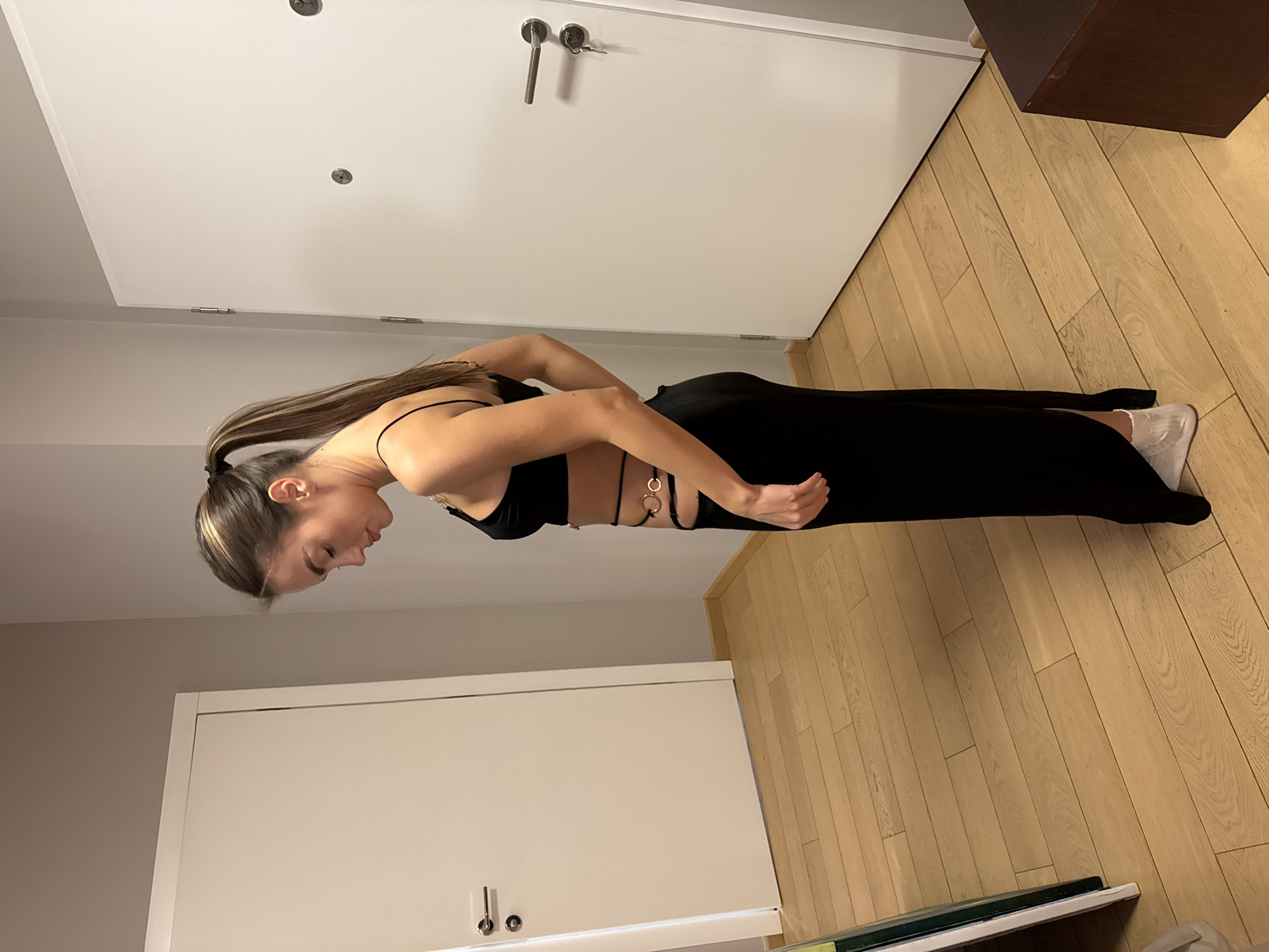 A woman with medium-length brown hair is standing indoors. She is wearing a black two-piece outfit with ring details around the midriff and white sneakers. The room has wooden floors. There are white doors visible in the background, one with a silver handle. The location is Warsaw, Poland.