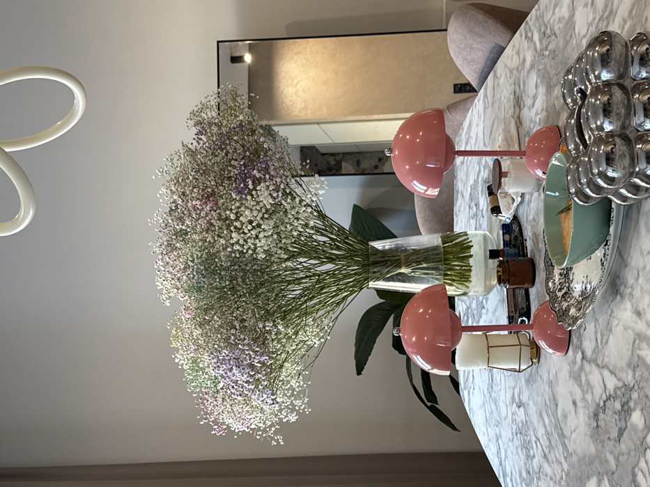 A lush bouquet of baby's breath dominates the image, displayed in a clear vase. Adjacent to the flowers, a set of two pink mushroom lamps with white candle holders sits on a marble surface. A decorative, tiered silver bowl holding a pastel green dish adds a touch of elegance. A mirror with a black frame is visible in the background, reflecting light and adding depth to the scene. The overall composition presents a stylish and modern interior setting.