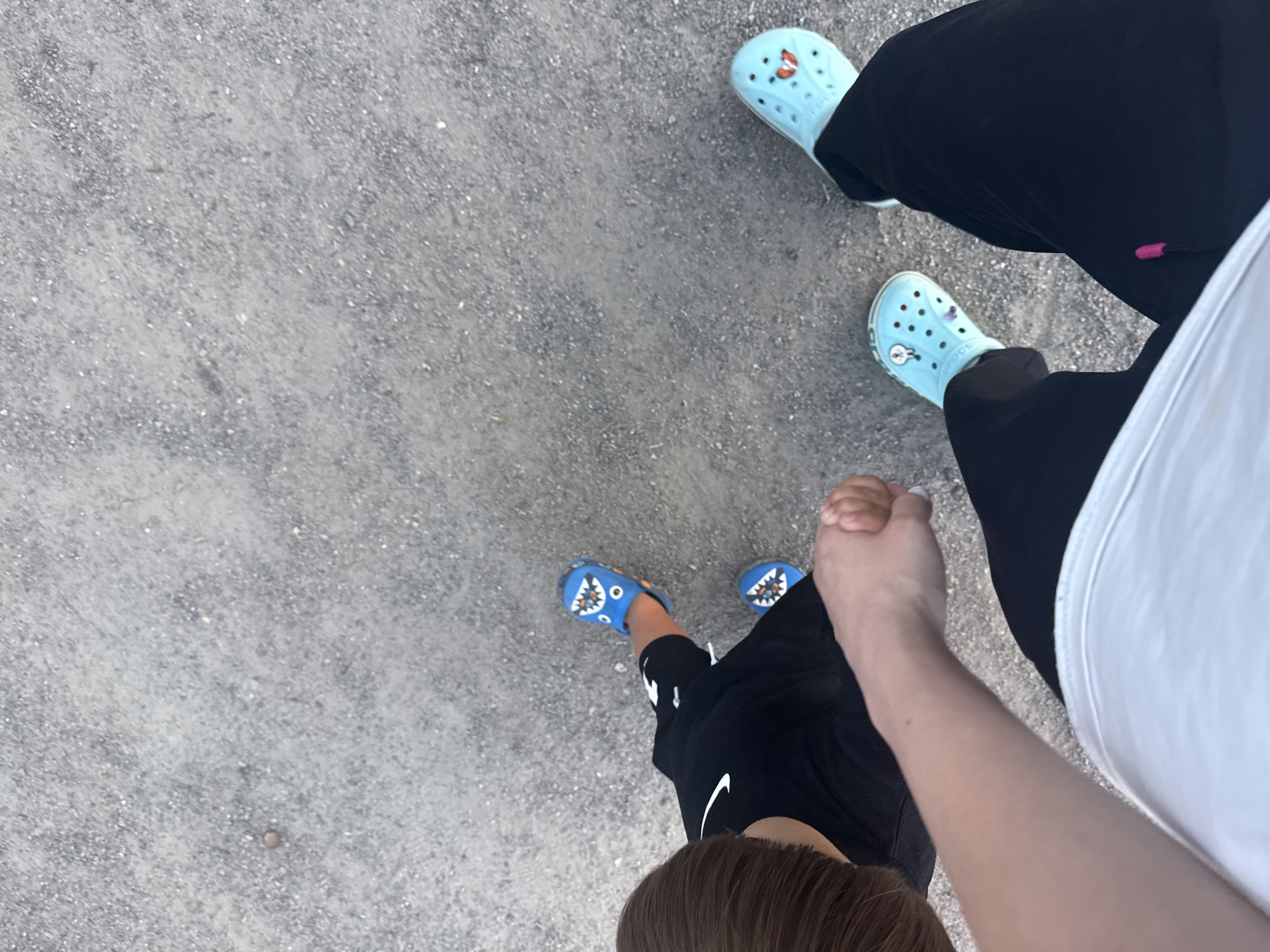 The image captures a high-angle, top-down view of two people's legs and feet walking on a gravel-covered surface. The person in the foreground, on the right, wears black pants and light blue Crocs with a cartoonish red face design. The person on the left, appearing to be a child, wears black shorts and blue Crocs featuring a shark design. The gravel appears to be a common outdoor surface, possibly a park or a walking path, common in Dnipro, Ukraine. The lighting suggests it is daytime. The context of the image is a casual walk, with the two individuals' legs in close proximity, suggesting they are together.