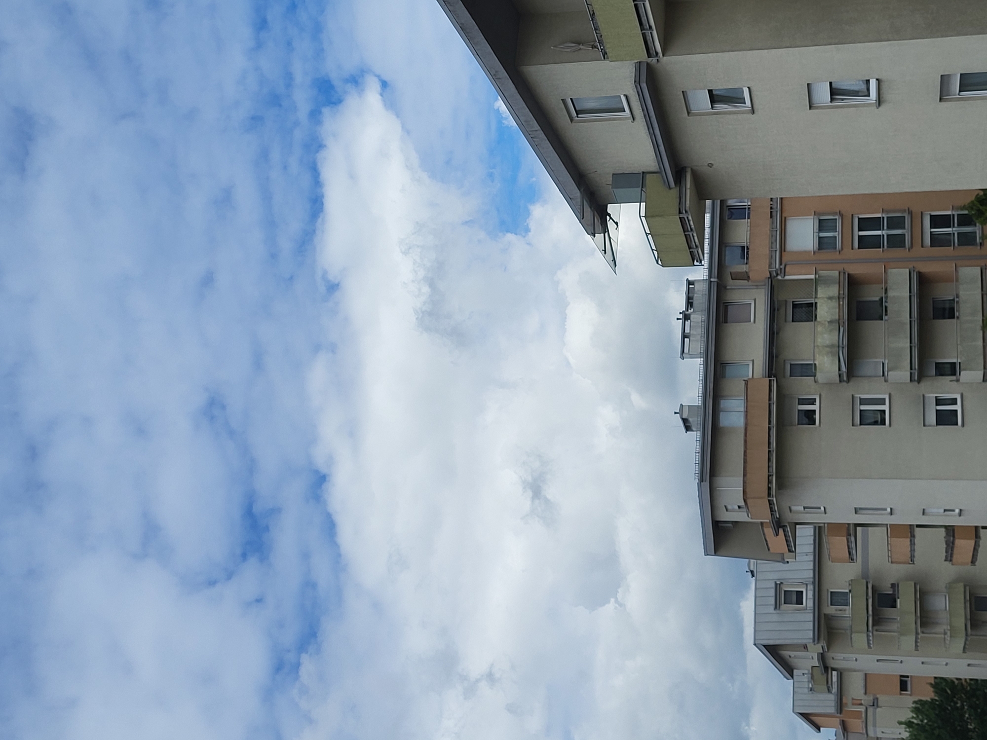 The image shows an outdoor scene with a cloudy sky and several apartment buildings. The buildings are multi-story structures with balconies and windows. The sky is filled with a mixture of white and gray clouds, with patches of blue sky visible. The buildings appear to be modern in design, with light-colored facades and some accent colors on the balconies. The overall impression is of a residential area on a partly cloudy day.