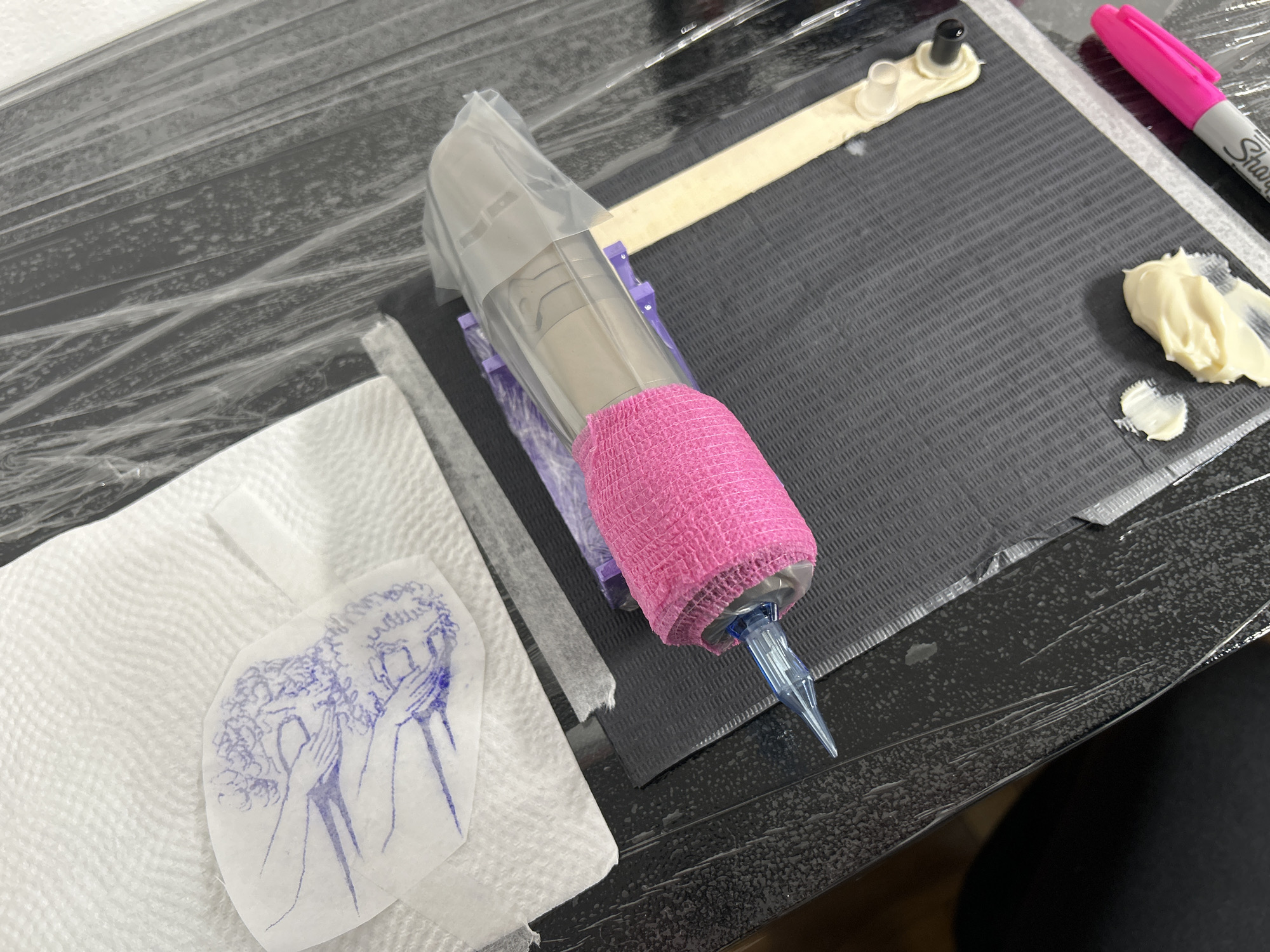 A tattoo artist's station is depicted, covered in clear plastic sheeting. On the left, a white paper towel holds a stencil of a person with their face in their hands. To the right of the stencil, a tattoo machine is wrapped in clear plastic, with a pink bandage-like material around the grip. The needle is visible. Next to the machine on the right is a black ribbed mat. A light-colored disposable tray with two small wells, one empty and the other filled with a black substance, lies on the mat. Next to the tray, a pink Sharpie marker rests on the mat, and to the marker's left, there is a dollop of a white creamy substance. The table on which the mat rests has a black finish with a visible wood grain texture.
There is no information to suggest the location in the city of Buenos Aires, Argentina.