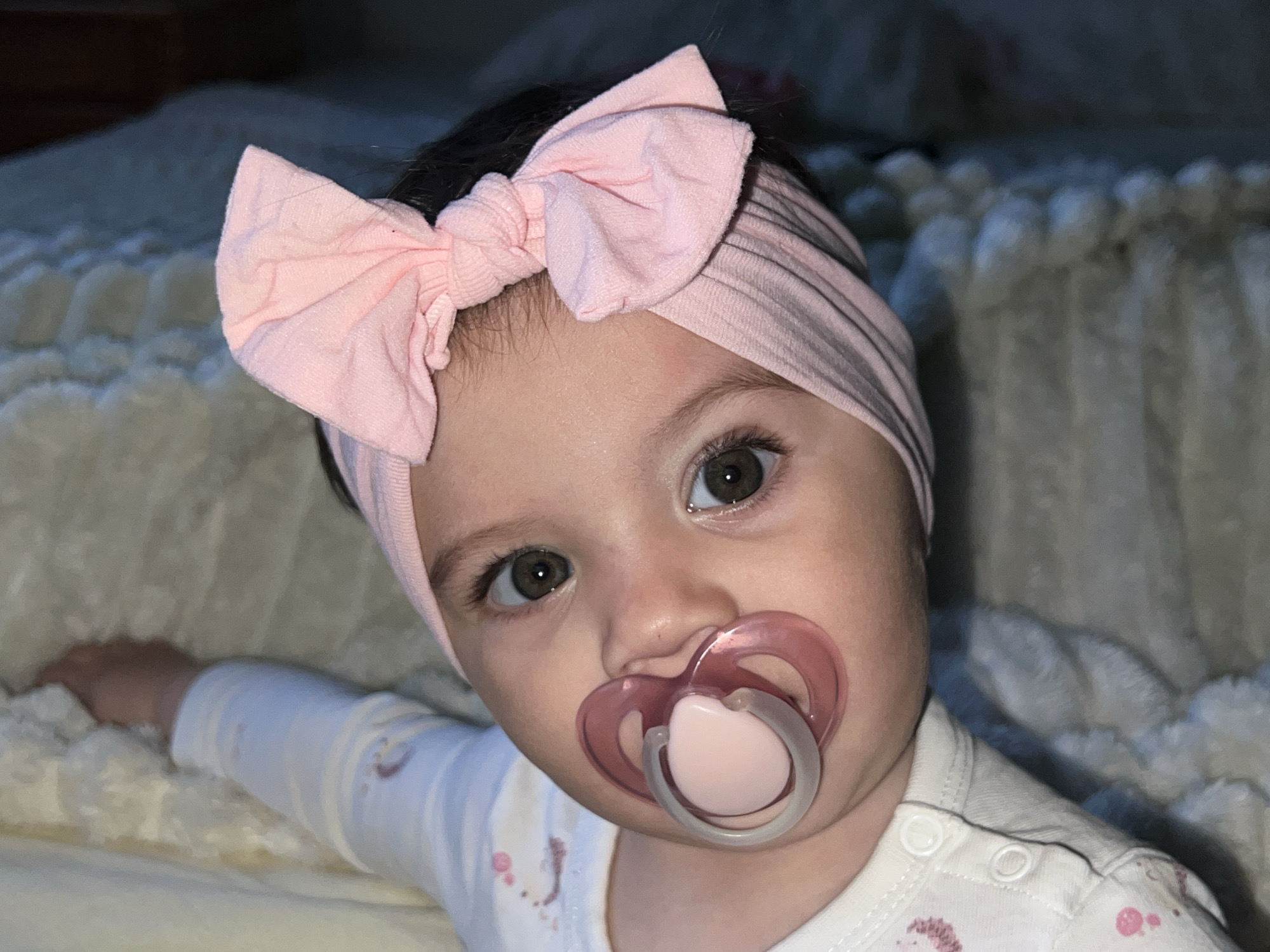 An infant is depicted in a close-up, chest-up portrait within an indoor setting in Elena, Bulgaria. The subject has dark hair, dark eyes, and a fair complexion, wearing a light pink ribbed headband with a prominent bow on top. A pink and translucent pacifier is present in the infant's mouth. The infant is dressed in a white long-sleeved garment featuring subtle light pink patterns. The right arm is extended, resting on a light-colored, textured surface, likely bedding, which also forms the out-of-focus background. The ambient lighting is dim.