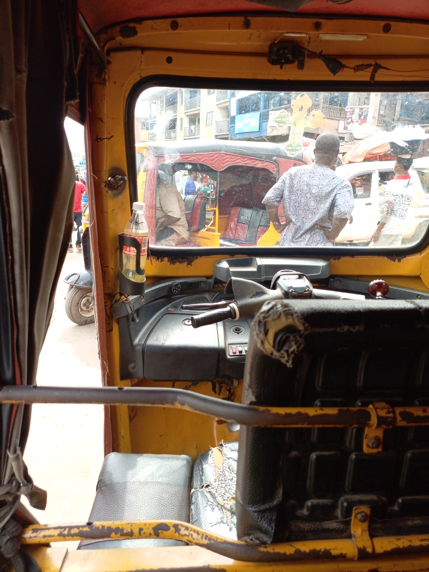 The image captures a street scene in Onitsha, Nigeria, from the perspective of a passenger inside a yellow tricycle taxi (Keke Napep). The viewpoint is from the rear seat, looking forward through the windshield and out onto the road.
In the foreground, the interior of the Keke Napep is visible, showing its yellow frame, a black dashboard with red buttons, and a worn, torn black steering handlebar. A black passenger seat, also showing wear, is positioned to the right. On the left side of the vehicle's frame, a plastic bottle containing a yellowish liquid (possibly oil or fuel) is held in a black holder; the label "BIG COLA" is partially discernible on the bottle. Brown curtains are drawn on the left side.
Through the windshield, a busy urban setting unfolds. A man with a short haircut, wearing a light-colored patterned shirt, stands with his back to the viewer in the midground, facing away from the taxi. To his right, another person in a white shirt with a large, square, QR code-like graphic and the word "TRADE" or "TRADERS" visible on the back, also stands facing away. In front of the Keke Napep, another tricycle taxi with a red canopy is visible. Further left, a man in a red t-shirt can be seen standing or walking.
The background features multi-story buildings with balconies, some painted blue or grey. A prominent, large yellow wooden structure, possibly a stylized cross or abstract sculpture, stands between the buildings. The activity suggests a stationary vehicle, possibly in traffic or parked, amidst pedestrians and other vehicles. The scene is set during daytime, under what appears to be overcast or diffused natural light. The interior of the Keke Napep exhibits signs of age and use, with noticeable wear and rust on the frame and upholstery.