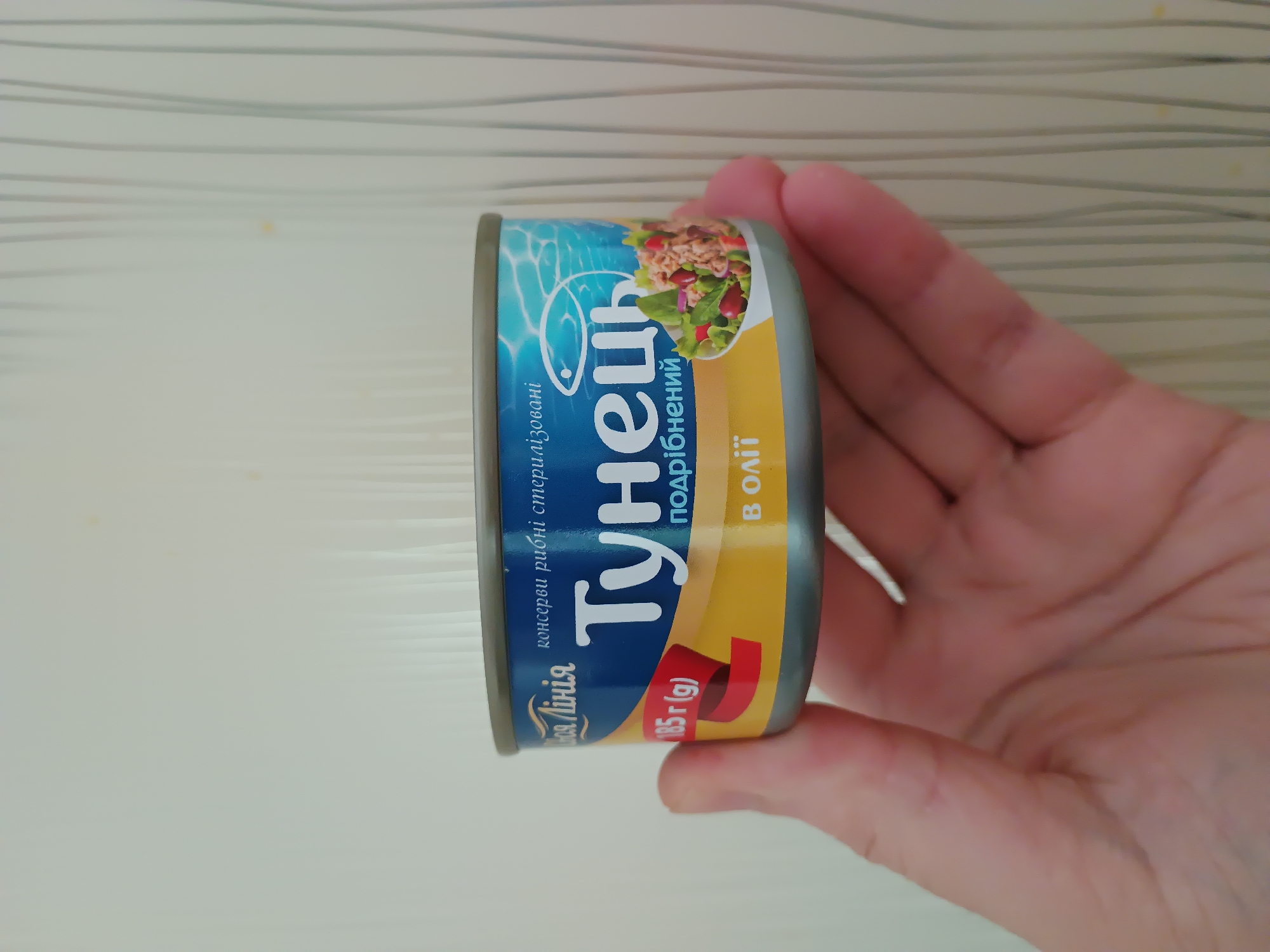 A hand holds a can of tuna, with the can label prominently visible. The can is blue and yellow, with text in Ukrainian. The text on the label includes "Тунець" (Tuna), "подрібнений" (chopped), and "в олії" (in oil). There is also a red ribbon with "185 г (g)" printed on it. The background is a pale, textured wall with vertical lines. The image was taken in Zaporizhzhia, Ukraine.