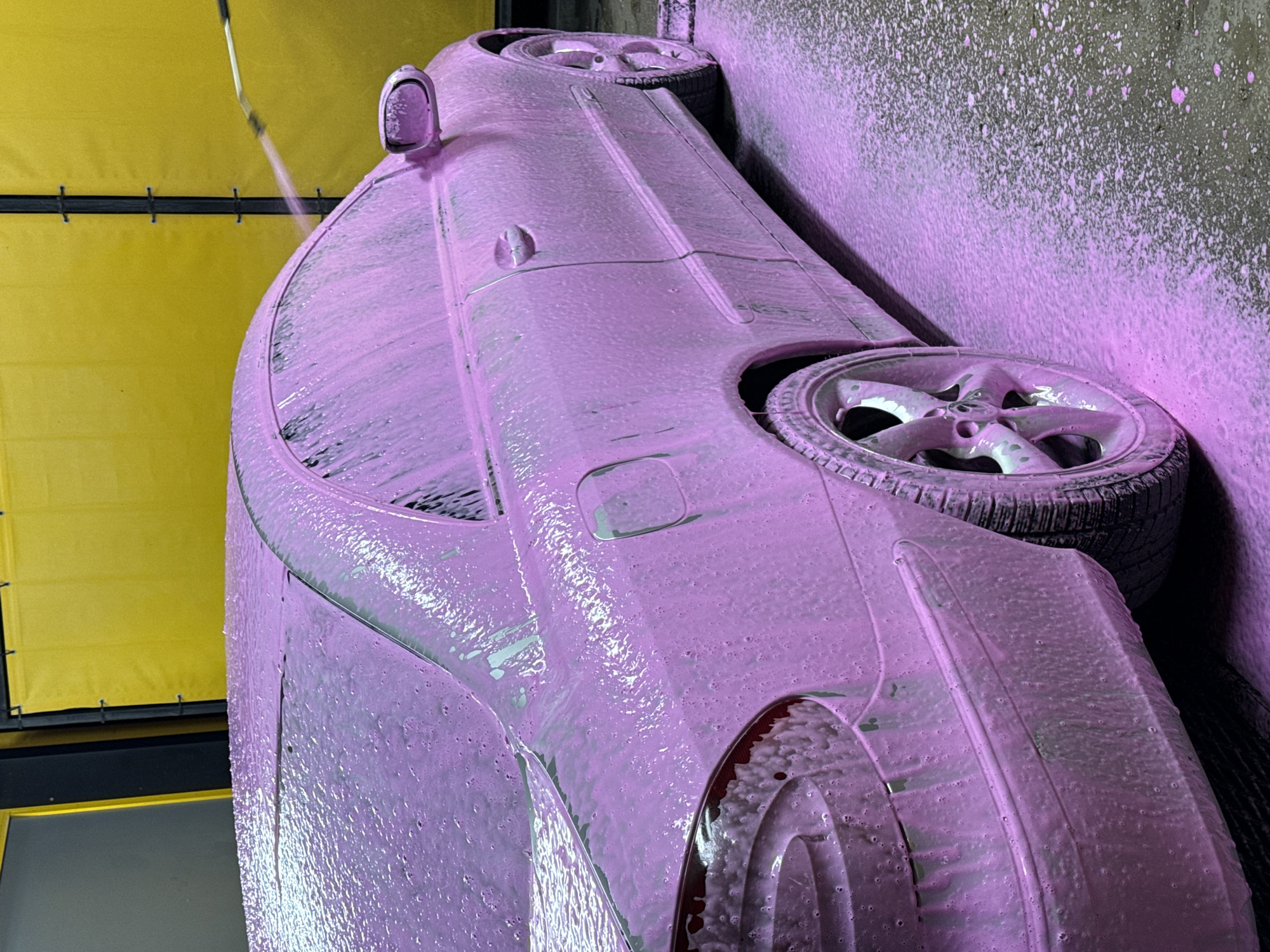 A pink-foamed car is being washed in a car wash. The car is a Mercedes-Benz CLK. The background consists of a yellow wall and a black frame. The scene appears to be taking place in a car wash in Pleven, Bulgaria.