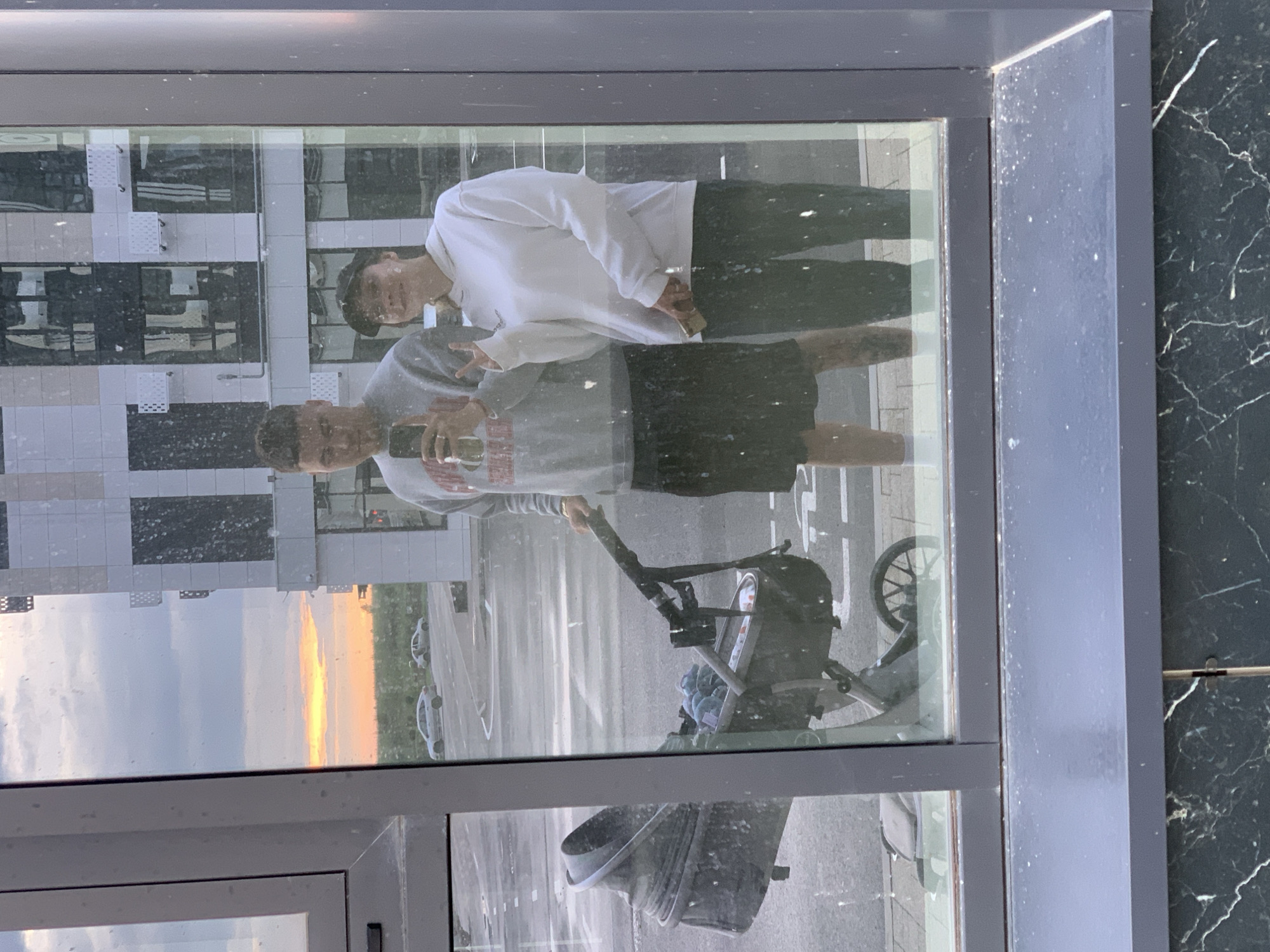 A mirror selfie captures a young couple and a stroller against a city backdrop. The man, wearing a grey sweatshirt and black shorts, holds a smartphone. The woman, in a white hoodie and black pants, smiles and makes a peace sign. A grey stroller with a baby is positioned to the left. Reflected in the glass are a modern building and a sunset sky. The scene is set in Stavropol, Russia.