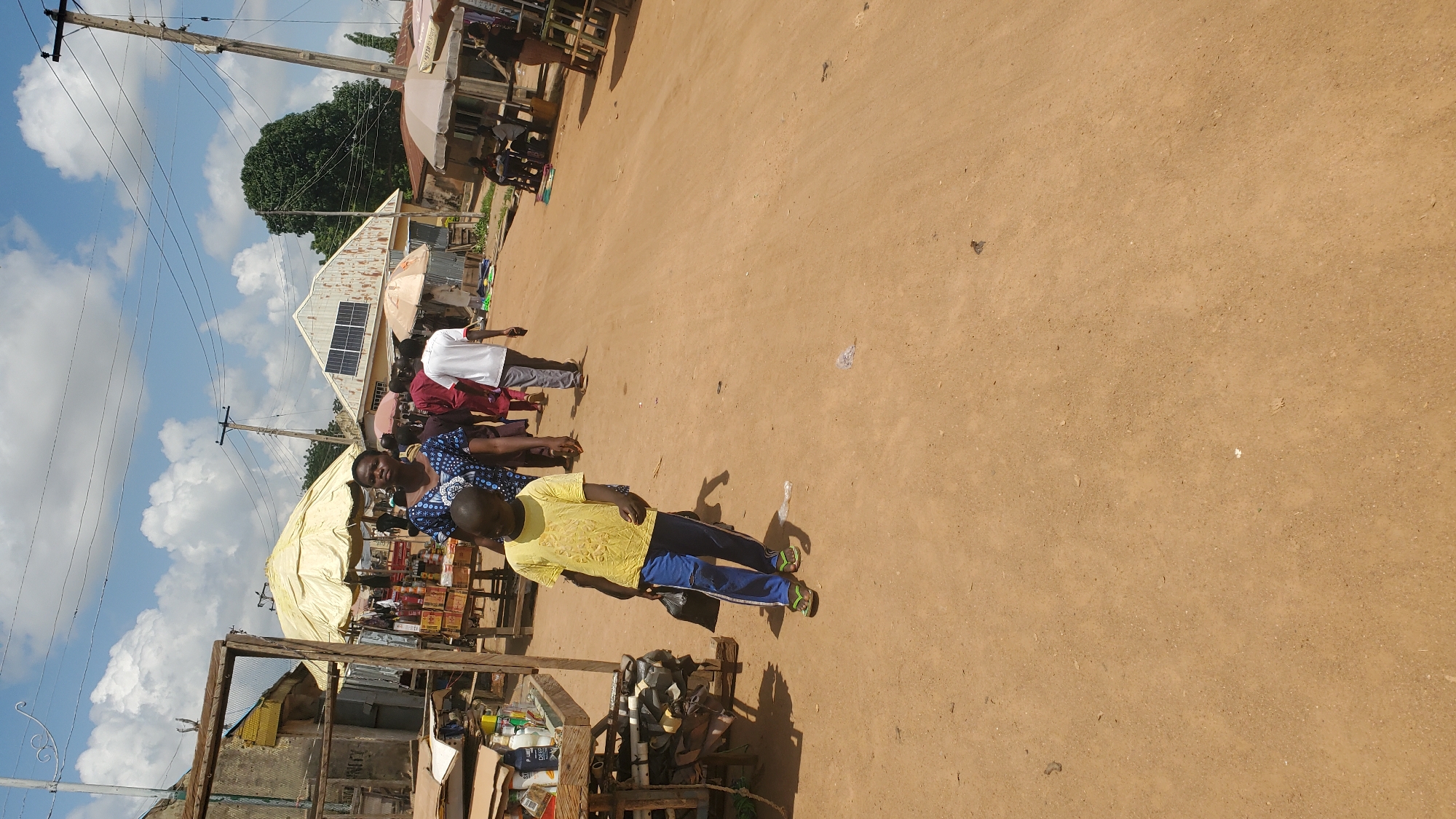 This is a medium shot, eye-level, outdoor photograph taken on a sunny day in Jalingo, Nigeria. The scene depicts a dirt road with various market stalls and people. In the foreground, a young boy in a yellow t-shirt and blue pants walks toward the camera, holding a black plastic bag. Behind him, a woman wearing a blue and white patterned dress is walking, along with other people in the background. Market stalls with umbrellas and shelves displaying goods are visible on either side of the road. In the middle ground, a building with solar panels on its roof stands out. Utility poles and power lines are present throughout the scene. The ground is covered in reddish-brown dirt, and there are some scattered pieces of trash on the road.