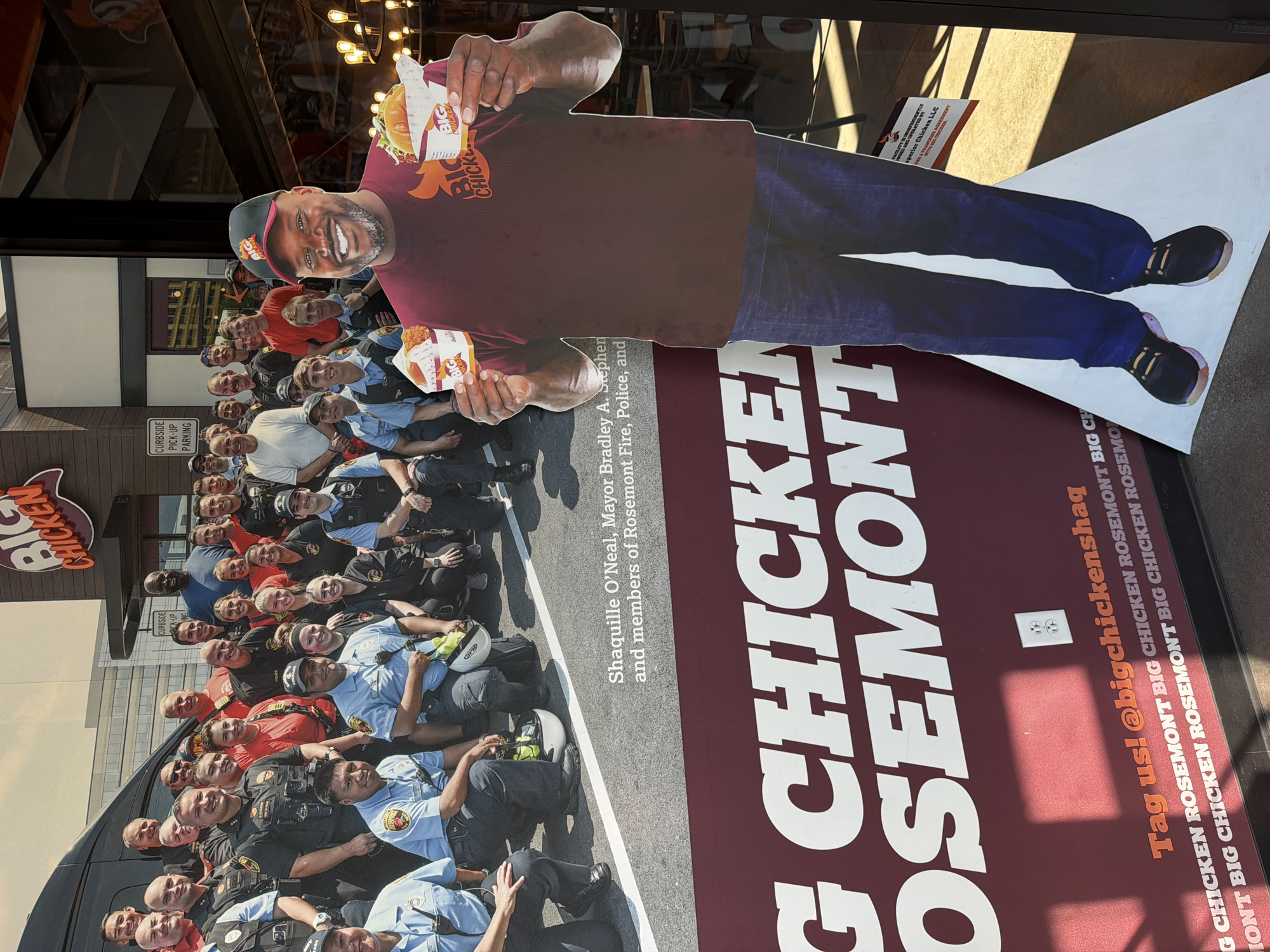 A cardboard cutout of Shaquille O'Neal stands in front of a poster advertising Big Chicken Rosemont, implying that this establishment is located in Rosemont, USA. The poster displays a group photograph with the text "Shaquille O'Neal, Mayor Bradley A. Stephen, and members of Rosemont Fire, Police, and," suggesting the photo features members of local fire and police services. The poster contains large text that reads "BIG CHICKEN ROSEMONT" and includes the phrase "Tag us! @bigchickenshaq" on the lower right of the poster. An electrical outlet is visible on the poster. The Big Chicken Rosemont logo is displayed in the top left of the group photograph. The scene is located on a sidewalk outside a Big Chicken Rosemont restaurant, as evidenced by the restaurant's exterior being reflected in the glass where the cutout is situated.