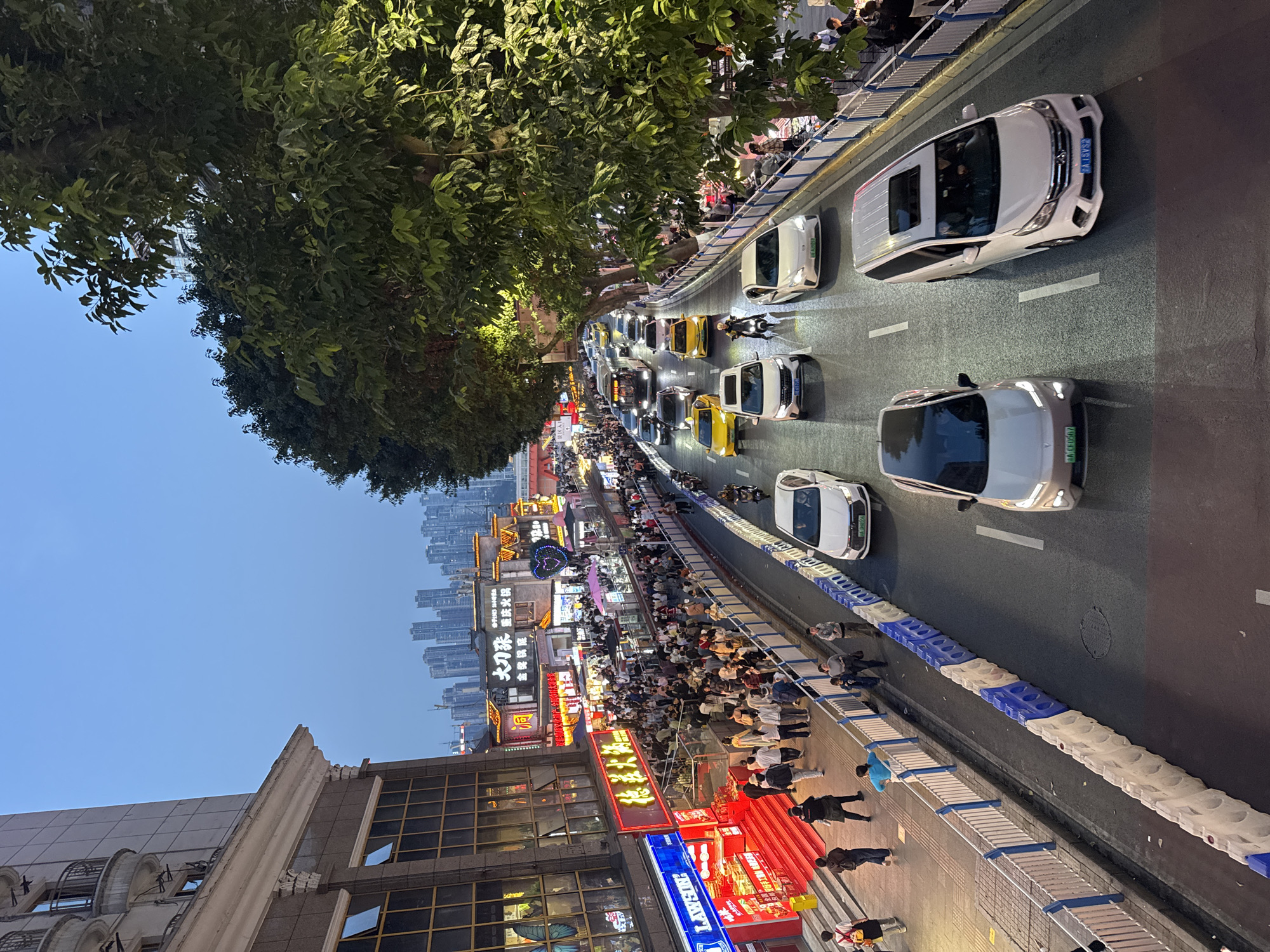 An evening view in Yuzhong District, China, shows a street scene with several cars on a multi-lane road separated from a pedestrian walkway by a blue and white barrier. A large tree overhangs the street. The pedestrian area is crowded with people walking and standing, adjacent to buildings with brightly lit storefronts featuring Chinese characters. Further in the background, the cityscape shows numerous tall buildings under a pale blue sky.