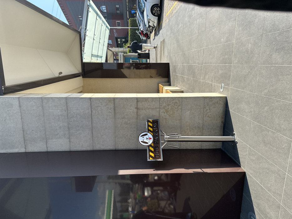 The image depicts an outdoor urban environment in Metamorfosi, Greece, under bright, clear daylight. The scene features modern architecture with tiled pavement, concrete walls, and glass elements.
In the foreground, a "DOG PARKING" sign is prominently displayed on a metallic pole. The sign has a white dog's head logo and yellow-and-black hazard stripes on a dark background, with the words "DOG PARKING" written vertically. This sign is situated next to a textured grey concrete wall. Below the sign, a large, dark, reflective surface, possibly a window or polished facade, mirrors parts of the surroundings.
Further back, a pedestrian walkway made of grey tiles extends into the distance. To the right of the walkway, several wooden slat benches are visible. In the background, a person wearing dark clothing and light-colored trousers stands near several parked cars on a paved street; a white car is clearly identifiable. Behind the street, various modern buildings with glass panels and darker facades are visible. A large, light-colored structure with an angular roof, possibly a canopy or part of a building entrance, occupies the left side of the frame.
The strong sunlight creates sharp shadows across the pavement and structures, indicating a sunny day, likely midday or early afternoon. No specific event or activity is underway beyond the presence of the individual in the background. The overall impression is that of a clean and contemporary urban space with designated amenities.