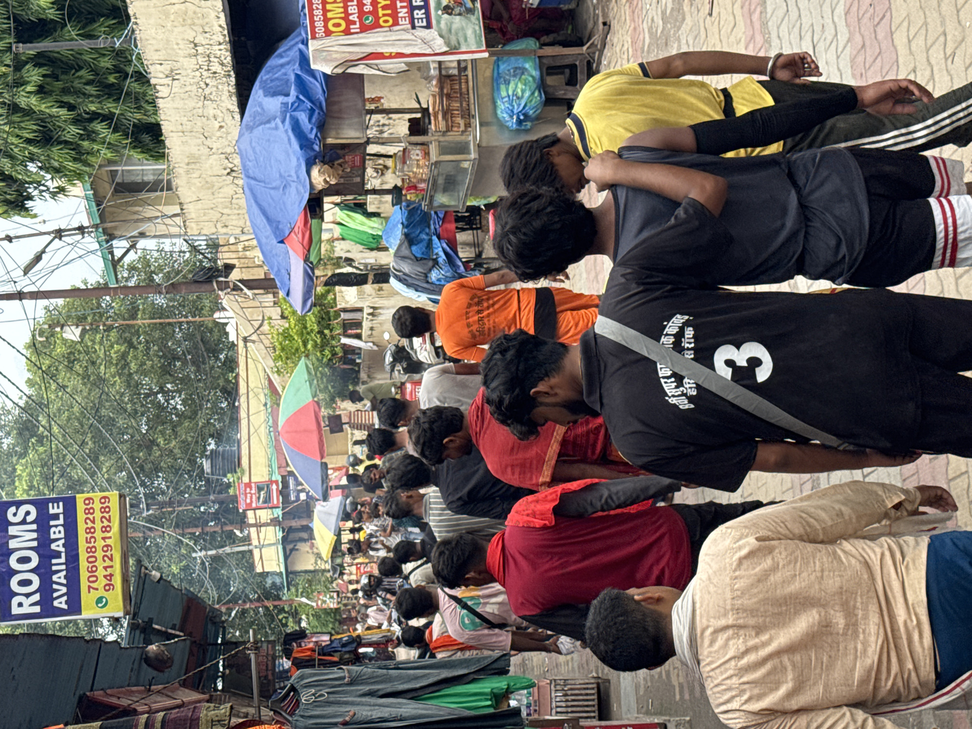 The image depicts a bustling street scene in Rishikesh, India, likely near the Lakshman Jhula bridge, a popular tourist area known for its ashrams, temples, and markets. In the foreground, a group of men are walking down a cobblestone street lined with shops. One man in a black t-shirt has Hindi text and the number "3" on his back. Another man next to him wears a grey tank top and black shorts with white stripes. A man in a yellow t-shirt walks behind them. The street is filled with more people, some wearing colorful clothing, and others in more muted tones.
Shops with goods displayed outside line the street. Some stalls have awnings and tarps, one with a large blue tarp partially covering goods. Several umbrellas, including a red, green, and yellow one and a large blue one, are visible, suggesting it might be a market area. In the background, buildings with balconies and windows can be seen, along with trees and utility poles with numerous wires. A sign hanging from above reads "ROOMS AVAILABLE" with phone numbers. The overall impression is one of a lively and crowded marketplace.