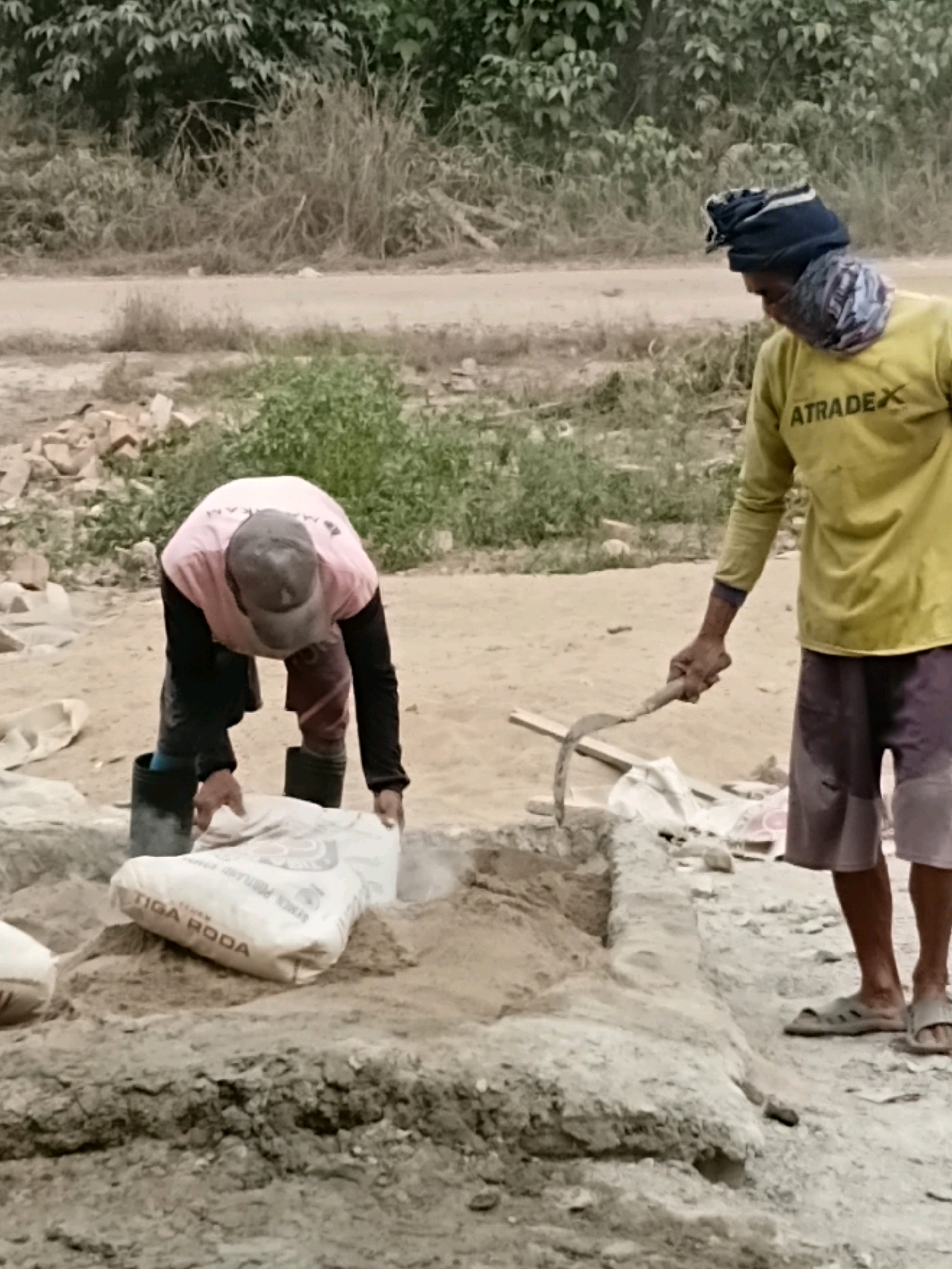 Two individuals are engaged in construction work in an outdoor, arid environment. The person on the left, wearing a pink long-sleeved shirt, dark pants, and black boots, is bent over a pile of sand, holding a large white bag that reads "TIGA RODA" and appears to be pouring its contents. The person on the right, dressed in a yellow long-sleeved shirt with the logo "ATRADE X" on it, dark shorts, and sandals, stands with a tool resembling a sickle in their right hand. The ground is sandy and dusty, with some piles of debris and sparse vegetation in the background. The overall setting suggests a construction site or a similar labor-intensive activity in a rural or semi-urban area of Margo Rukun, Indonesia.