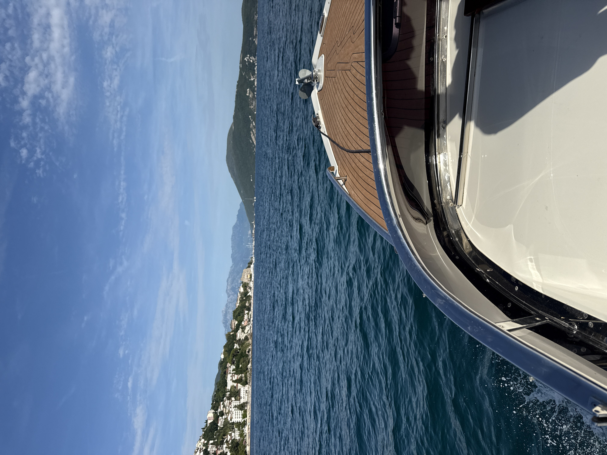 The image captures a first-person perspective from the bow of a moving boat, likely a motor yacht, on deep blue-green water under a clear blue sky with sparse white clouds. The boat's foredeck features a brown, horizontally striped surface, possibly faux teak, with a chrome anchor mechanism and a black line visible at the very front. Polished metallic rails and a white hull section are prominent in the foreground, with water spray indicating the boat's forward motion.
The scene depicts the coastal landscape of Herceg Novi, Montenegro. To the left, the city's densely built environment is visible, with numerous white and light-colored buildings ascending the steep green hillsides. Mountains rise in the distant background beyond the urban sprawl. Further along the coastline, hints of a harbor or marina with boat masts are discernible. To the right, a large, prominent, dark green, rounded landmass, identified as part of the Lustica Peninsula, extends into the water, with scattered buildings along its base. The bright sunlight casts distinct shadows on the boat's white deck.