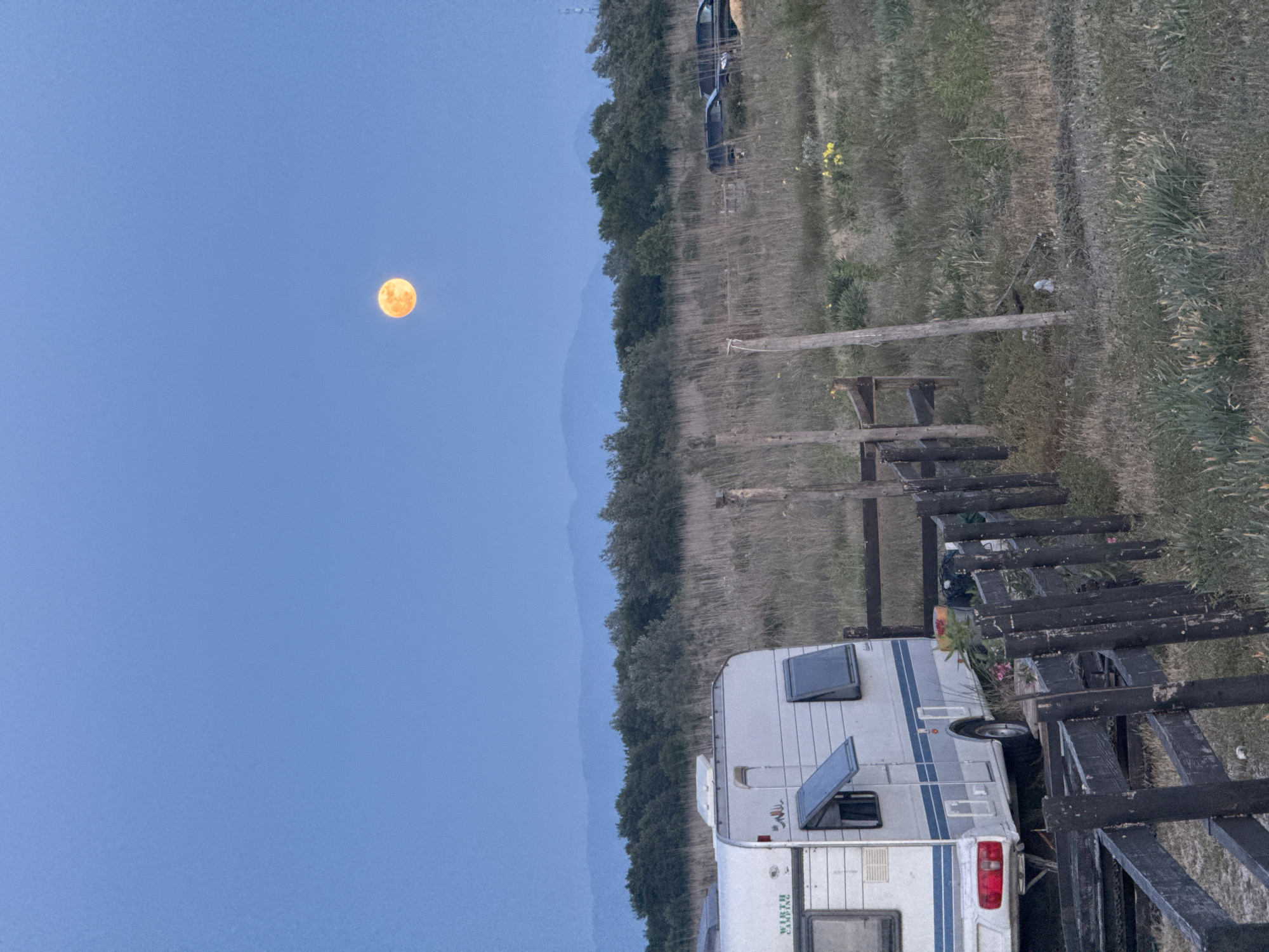Observed in Gornji Štoj - Shtoji i Epërm, Montenegro, is a twilight scene featuring a full moon positioned prominently in a clear, blue sky. Below the horizon lies a rural landscape encompassing a forested area. In the foreground, a white camper van is parked near a wooden fence. The terrain appears grassy and partially overgrown. A distant vehicle is also noticeable further back in the shot.