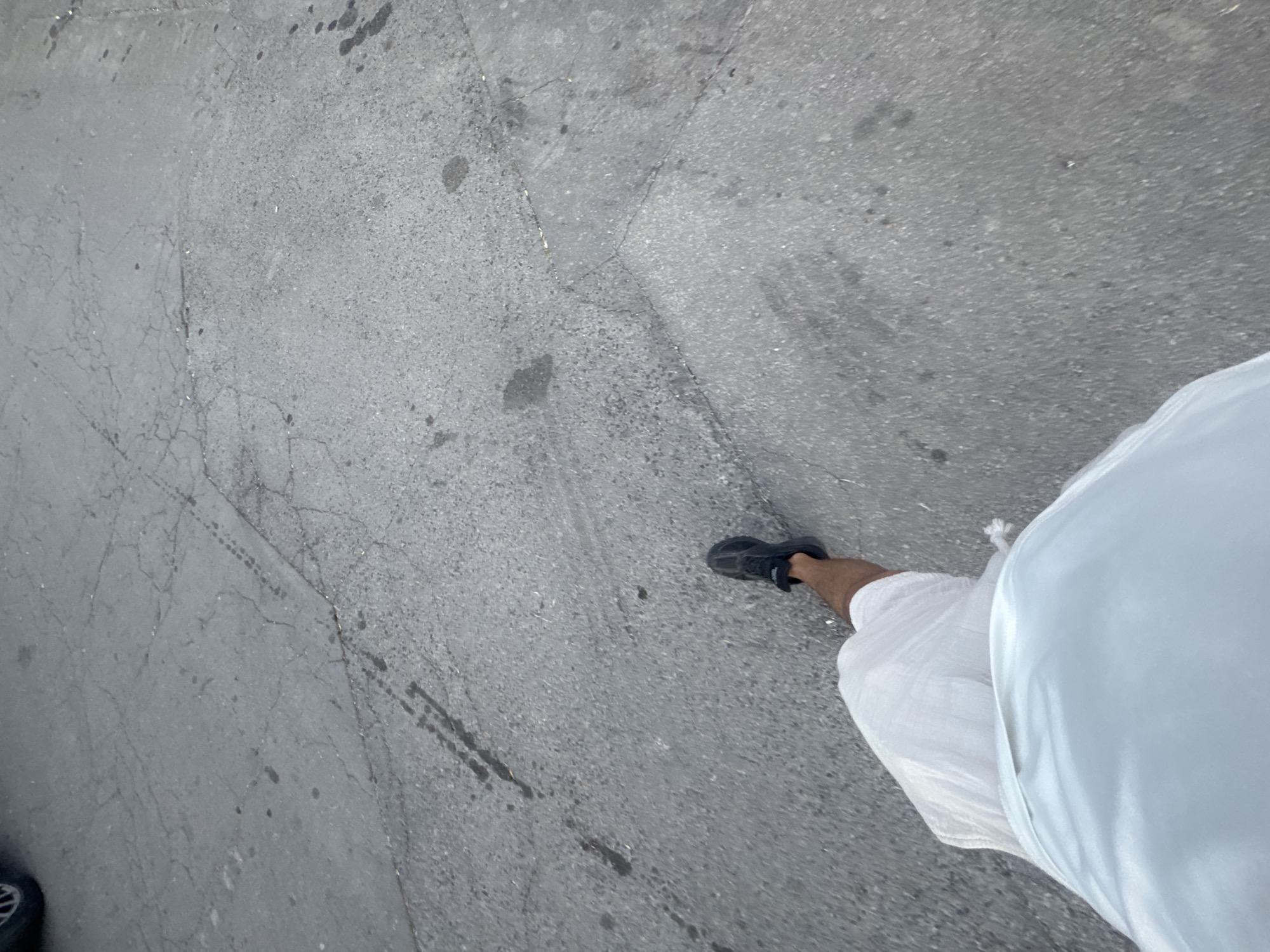 The image shows a low-angle, first-person perspective of a person walking on a cracked asphalt surface. The person is wearing white shorts and black sneakers. The environment appears to be an outdoor urban setting, likely a street or sidewalk, with some tire marks and dark stains on the asphalt. There are no other people or significant landmarks visible in the frame. The lighting suggests it might be daytime. The location is not specified in the image.