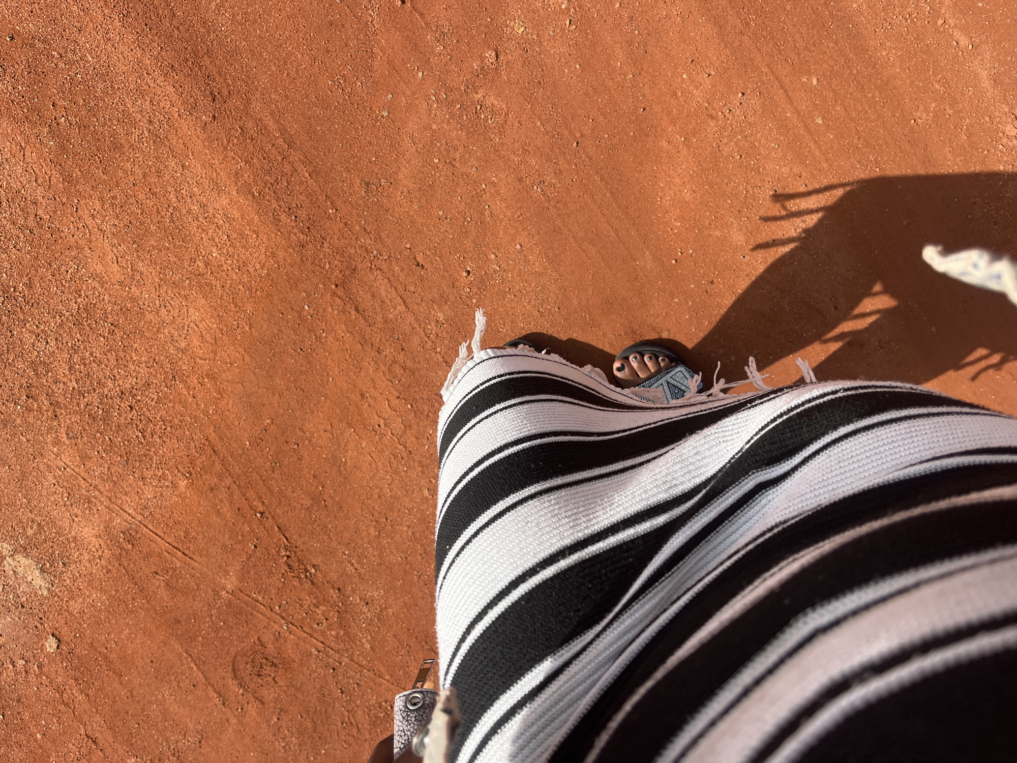 The image shows a close-up view from a high angle of a person's legs and feet on a red clay surface, likely a tennis court. The person is wearing a black and white striped skirt or dress, and a sandal on their left foot. The skirt has frayed edges. The ground is covered in fine red dust and small pebbles, with some faint tracks visible. A shadow is cast on the ground, indicating a sunny day. The scene appears to be outdoors, possibly in a recreational area or sports facility. The location is described as Bukuru, Nigeria.