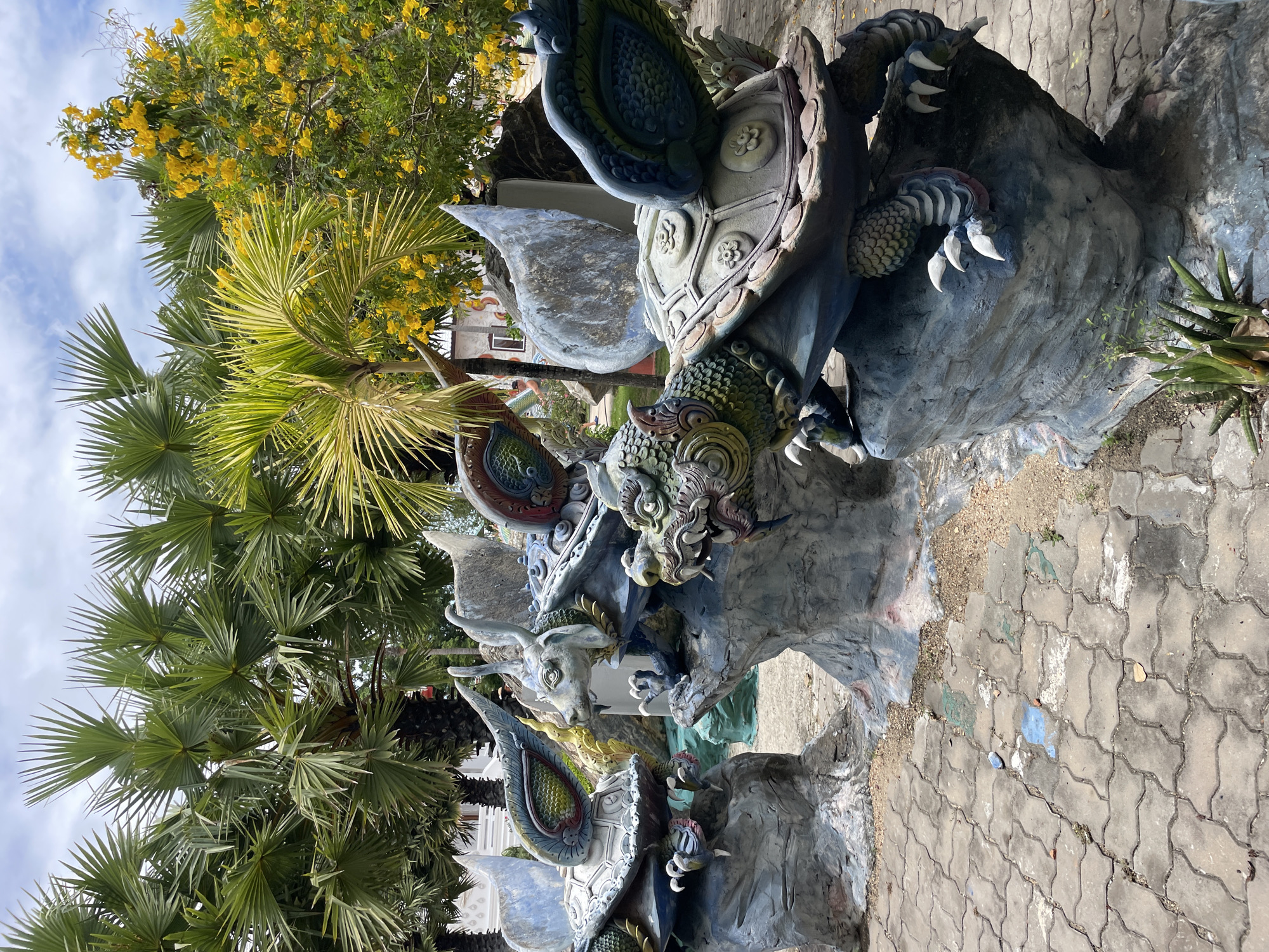 The image shows a courtyard with paved ground, likely in Bang Sare, Thailand. In the foreground, there are three large, weathered concrete sculptures of mythological creatures resembling turtles with dragon-like heads and wings. These sculptures are perched on rocky bases. Palm trees with large fronds and a tree with yellow flowers are visible in the background, suggesting a tropical environment. The sky is partly cloudy. The scene appears to be outdoors, possibly in a temple complex or a tourist attraction.