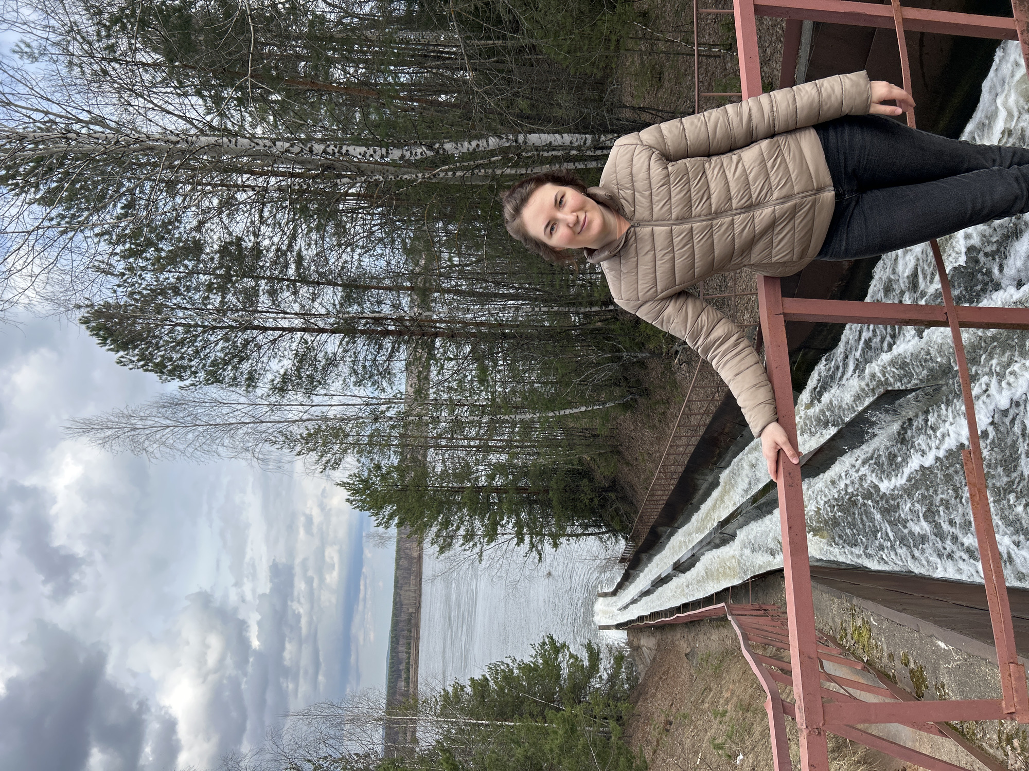 A woman is standing on a rusty red metal railing overlooking a waterfall. The waterfall consists of multiple tiers of water cascading down a concrete structure. In the background, there is a lake surrounded by trees. The sky is cloudy. The woman has brown hair and is wearing a beige jacket and dark jeans. She is smiling at the camera. The location is possibly near the city of Chernushka, Russia.