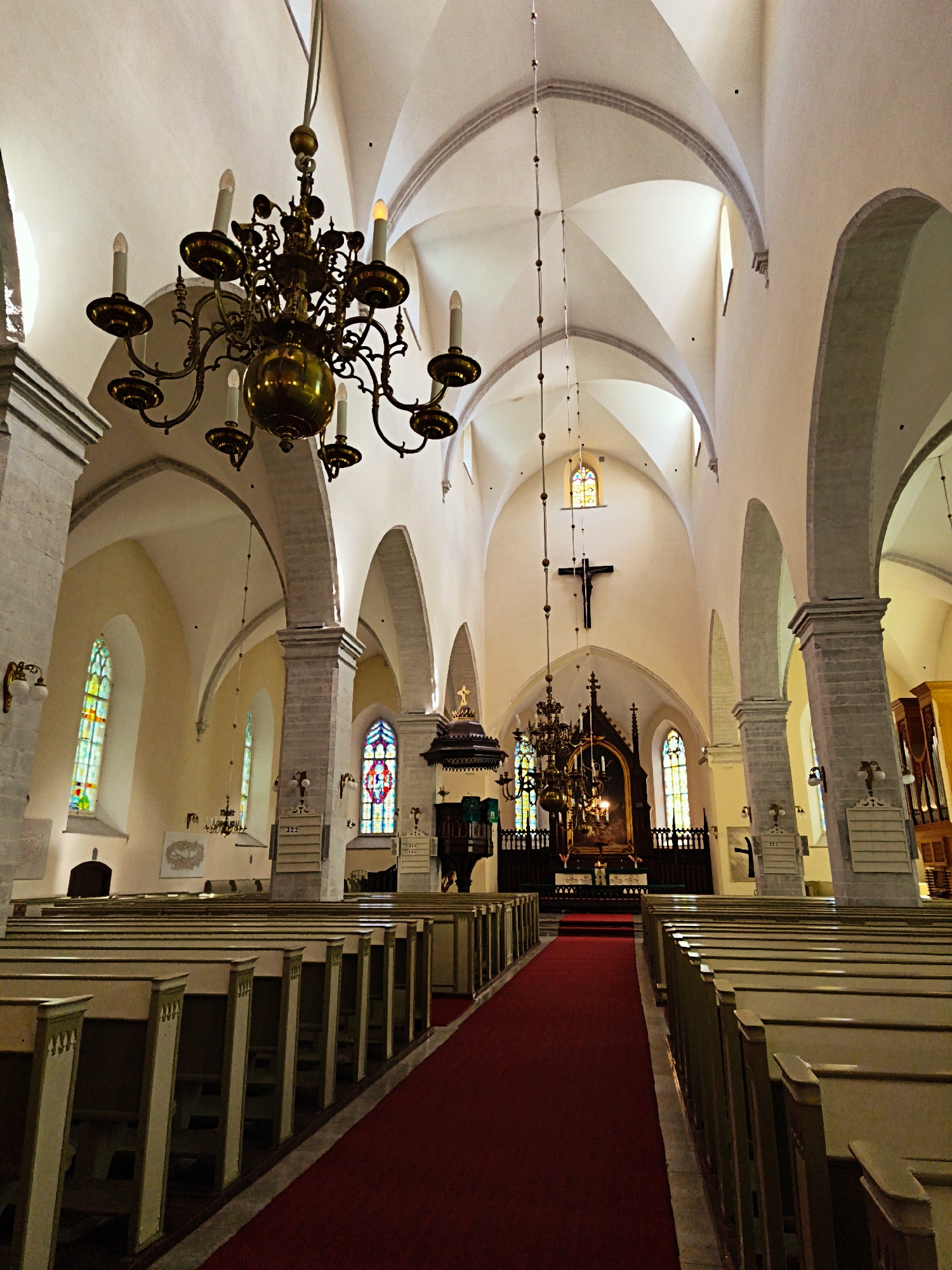 The interior of a large, ancient stone church in Tallinn, Estonia, is shown. The perspective is from the back of the nave, looking towards the altar. Pews line both sides of a red carpeted aisle that leads to the front. A large brass chandelier hangs from the arched, vaulted ceiling. Stained glass windows, featuring colorful geometric patterns, are visible on the walls. A crucifix hangs on the wall behind the altar, which is adorned with a painting and flanked by a pulpit. The architecture suggests a Gothic style, with ribbed vaults and pointed arches. The lighting is dim, with the primary illumination coming from the stained glass windows and some artificial lights. The church appears to be empty, with no people present.