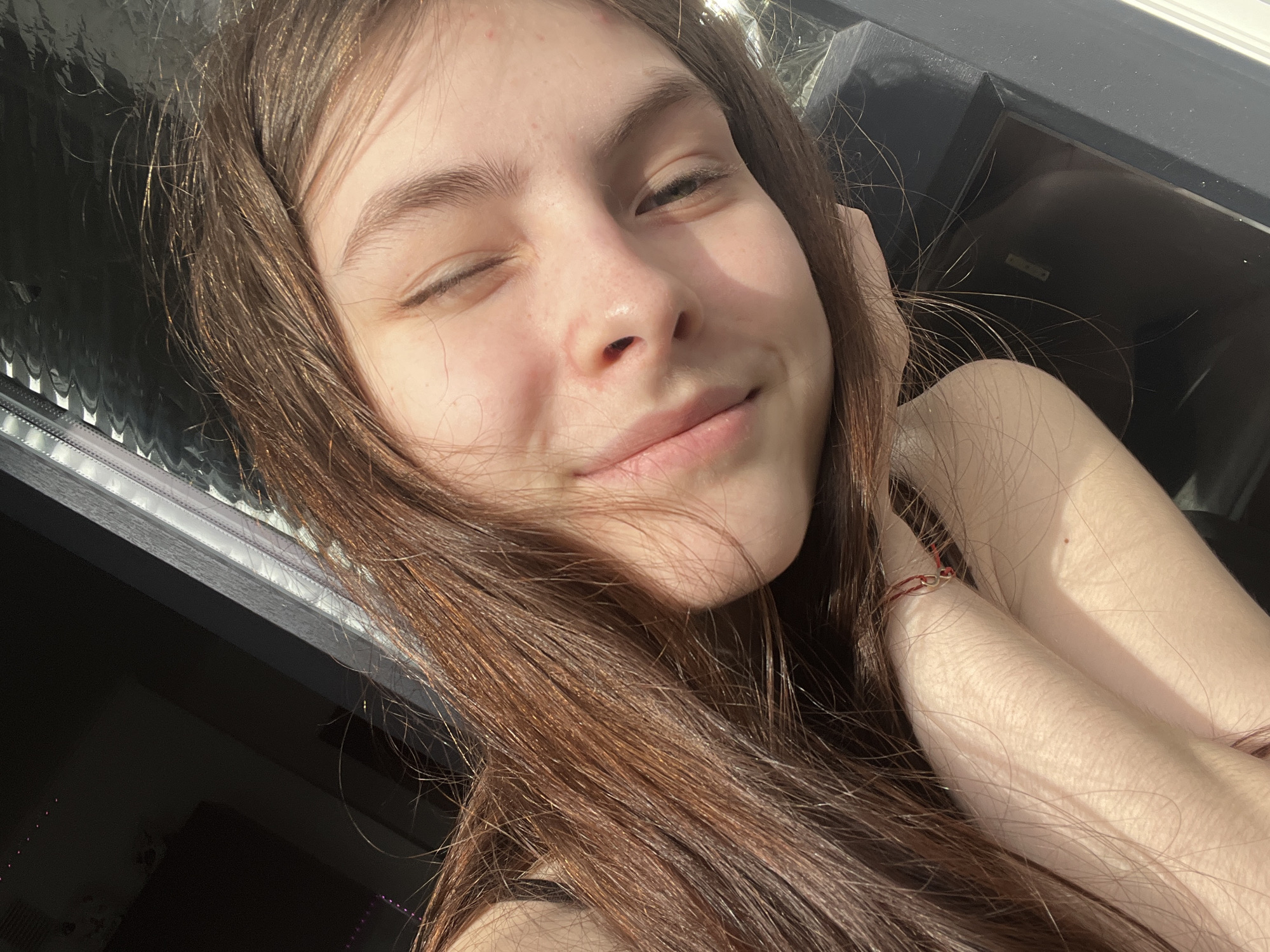 A fair-skinned, young woman with long, light brown hair takes a selfie. She is partially winking and smiling. Her right arm and hand are raised to the right side of her face. A red string bracelet is visible on her wrist. Behind her is a closed window with a black frame, and a blurry view of what appears to be city scenery. The location cannot be determined with the available information.