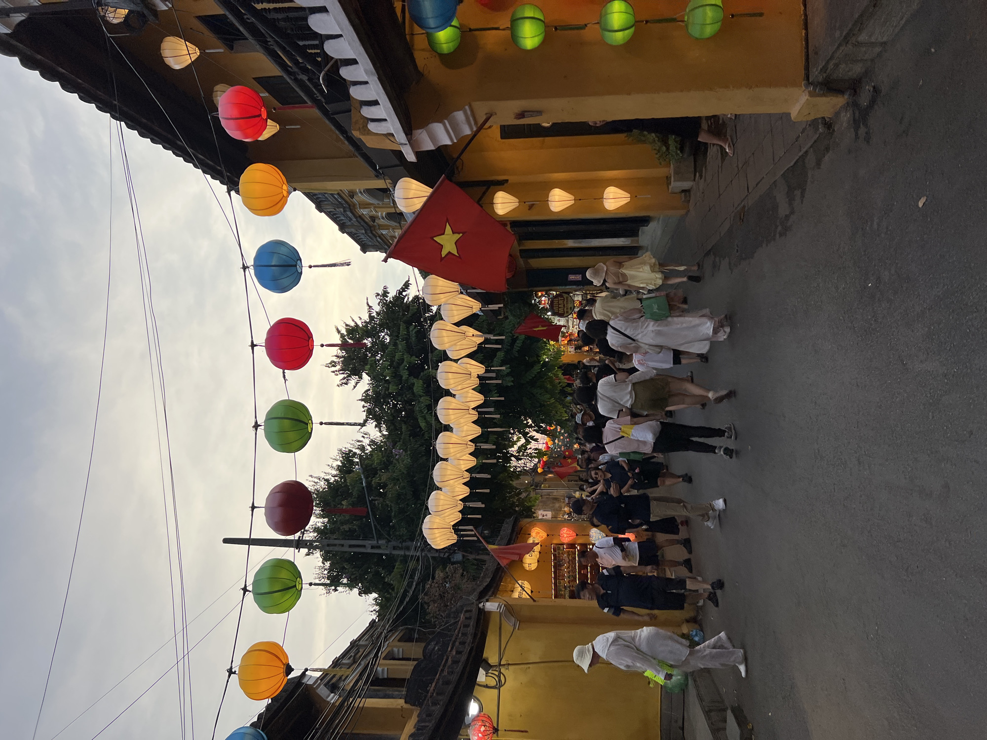 The image depicts a street scene in Hội An Ward, Vietnam, likely during the evening or dusk, given the lighting and the presence of illuminated lanterns. A diverse group of people are walking down a paved street lined with buildings adorned in yellow. Overhead, numerous colorful lanterns are strung across the street, creating a festive atmosphere. Several of these lanterns are lit, casting a warm glow. One prominent lantern is red and white with a star, resembling a Vietnamese flag. The buildings on either side of the street have traditional architecture with tiled roofs and wooden balconies. Some of the buildings appear to be shops or businesses, indicated by signage. The overall impression is one of a bustling tourist area, characterized by its vibrant decorations and pedestrian activity.