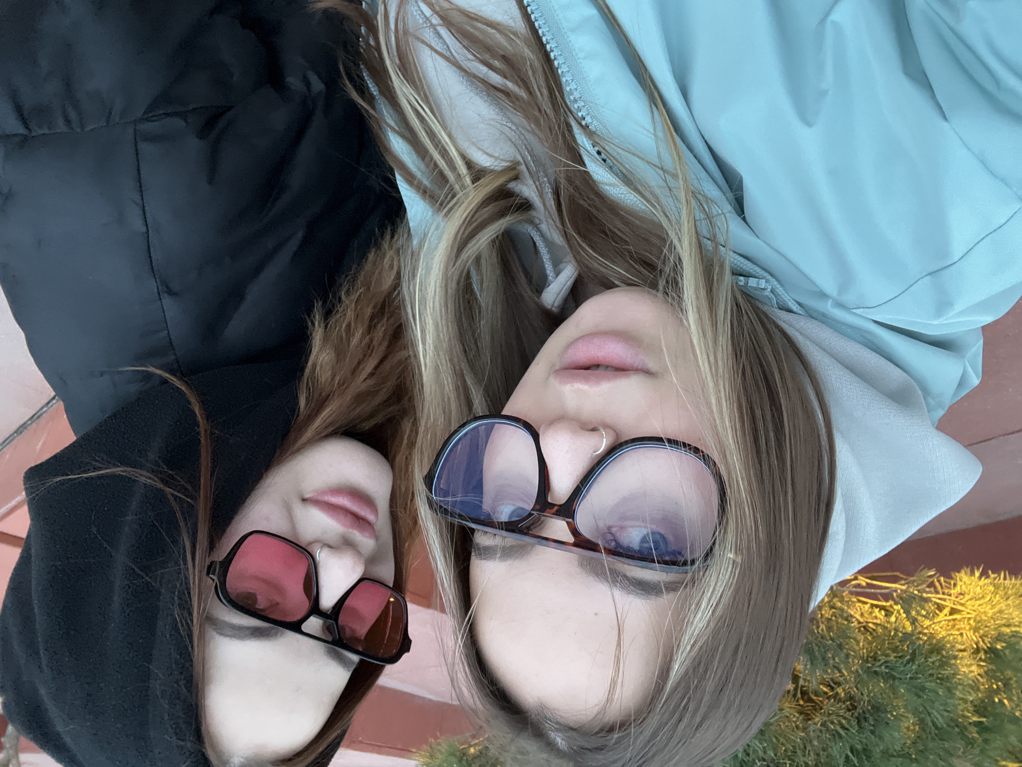 This close-up, overhead photograph captures two young women lying side-by-side in an outdoor setting, potentially a selfie or taken from above. The image is oriented vertically, showcasing their faces and upper torsos.
The woman on the left has dark brown hair, which frames her face. She is wearing a black hooded puffer jacket and stylish, square-framed sunglasses with red-tinted lenses. A silver hoop nose ring adorns her left nostril, and her lips are a soft pink.
To her right, the second woman has long, blonde and light brown streaked hair. She is dressed in a light blue hooded jacket or hoodie. She wears larger, square-framed glasses with a tortoiseshell pattern and clear lenses, which reflect the ambient light and obscure her eyes. She also has a silver hoop nose ring on her left nostril, and her lips are a similar soft pink hue.
Both women appear relaxed, with subtle, content expressions. The background behind them features a warm, reddish-brown surface, possibly a deck or pathway. On the far right, a patch of vibrant green foliage, likely coniferous, is brightly illuminated by golden sunlight, suggesting either early morning or late afternoon. The overall atmosphere is casual and serene. No text is visible in the media file. The location is Krasnodar, Russia.