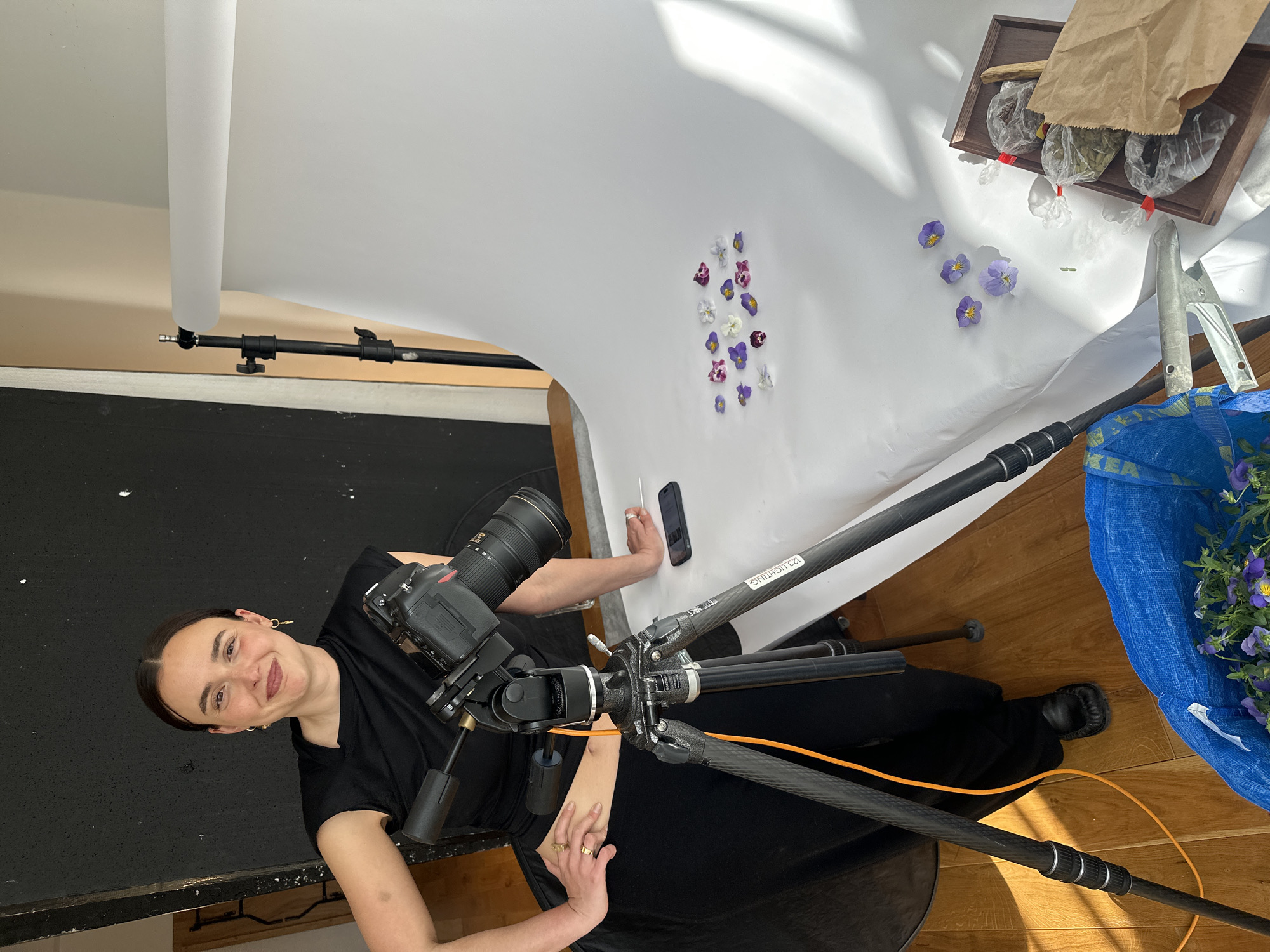 A female photographer is captured in an indoor setting, likely a photography studio, engaged in a still-life photoshoot. The image is taken from an elevated perspective, looking down at the setup and the smiling photographer.
The woman, with dark hair pulled back, wears a black short-sleeved top and has rings on her fingers and hoop earrings. She is looking towards the viewer with a friendly, subtle smile. Positioned beside her is a professional DSLR camera with a large lens, mounted on a sturdy, dark-colored tripod, possibly made of carbon fiber. An orange cable is visible wrapped around a tripod leg, which also bears the text "123 LIGHTING".
The primary subject of the photoshoot is arranged on a large white seamless paper background, which curves upwards to create a smooth sweep. Scattered across this white surface are numerous small pansy flowers in shades of purple, white, and pink. To the right, a wooden tray holds three clear plastic bags containing dried botanical items, possibly herbs or spices, along with a brown paper bag. A black smartphone and a silver stylus or tool are also resting on the white background near the photographer's hand.
On the wooden floor to the right, a blue IKEA Frakta bag, emblazoned with the text "IKEA", is overflowing with more pansy flowers. A large silver clamp is also visible on the floor. On the left side of the frame, a black light stand or boom arm supports the roll of white paper. The scene is well-lit, with natural light creating distinct shadows and highlights on the white background, suggesting it is daytime. A dark, possibly black, surface runs along the left edge of the image. The setup indicates a focused and creative environment.