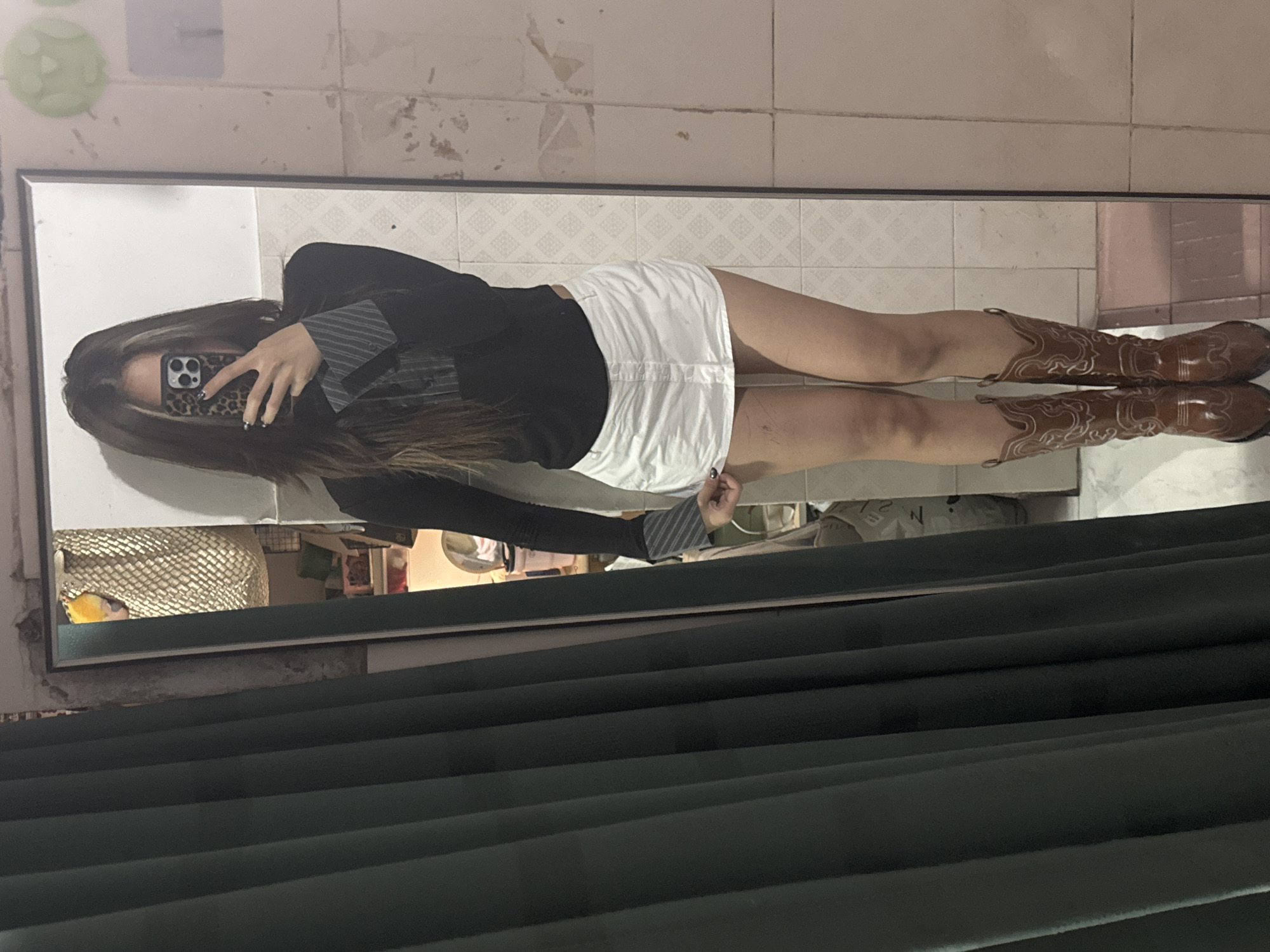 The image shows a woman taking a mirror selfie indoors. She is wearing a black blazer over a striped shirt, a white mini skirt, and brown cowboy boots. She is holding a phone with a leopard print case. The background reflection includes a tiled wall, a mirror with visible dust and smudges, a glimpse of a room with various items, and a window partially covered by dark green blinds. The location is likely in a residential setting in the city of Nan'an District, China, though this cannot be definitively confirmed from the image.