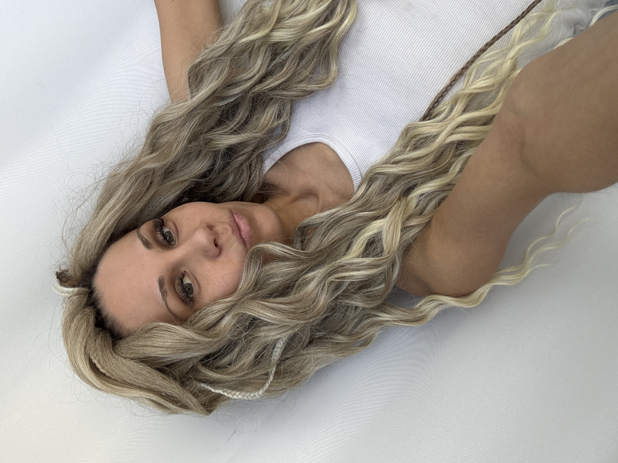 A woman is lying on a white surface. She is wearing a white ribbed tank top. Her long, wavy blonde hair is spread around her. Her skin tone is fair, and she has light brown eyes and noticeable eyelashes. Parts of her arms are visible on either side of her. The background is white. The image does not contain any information that could identify the location as the city of Rusanovo, Russia, or any other specific location.